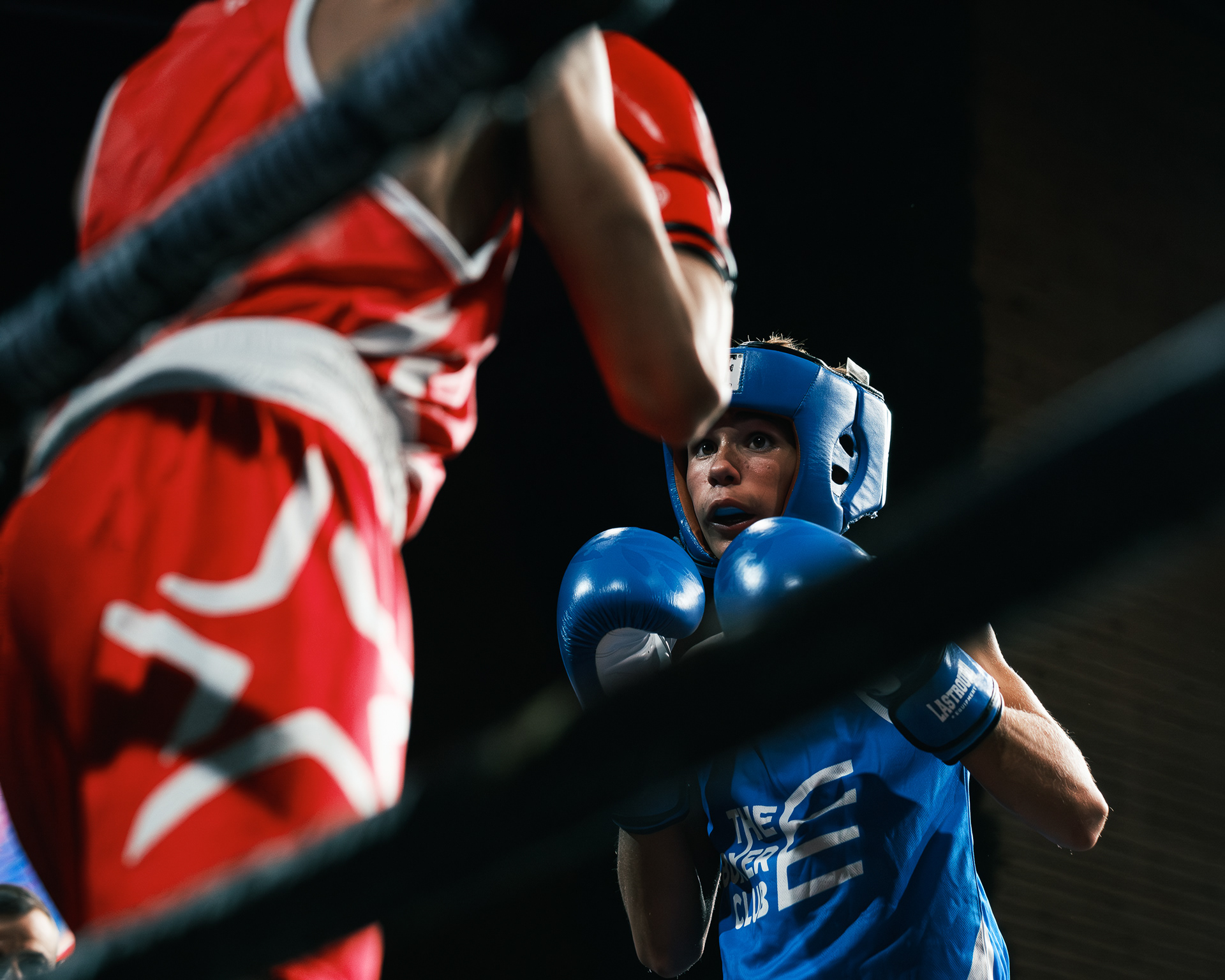 Boxeador con casco y guantes azules en guardia frente a un rival vestido de rojo, visto entre las cuerdas.