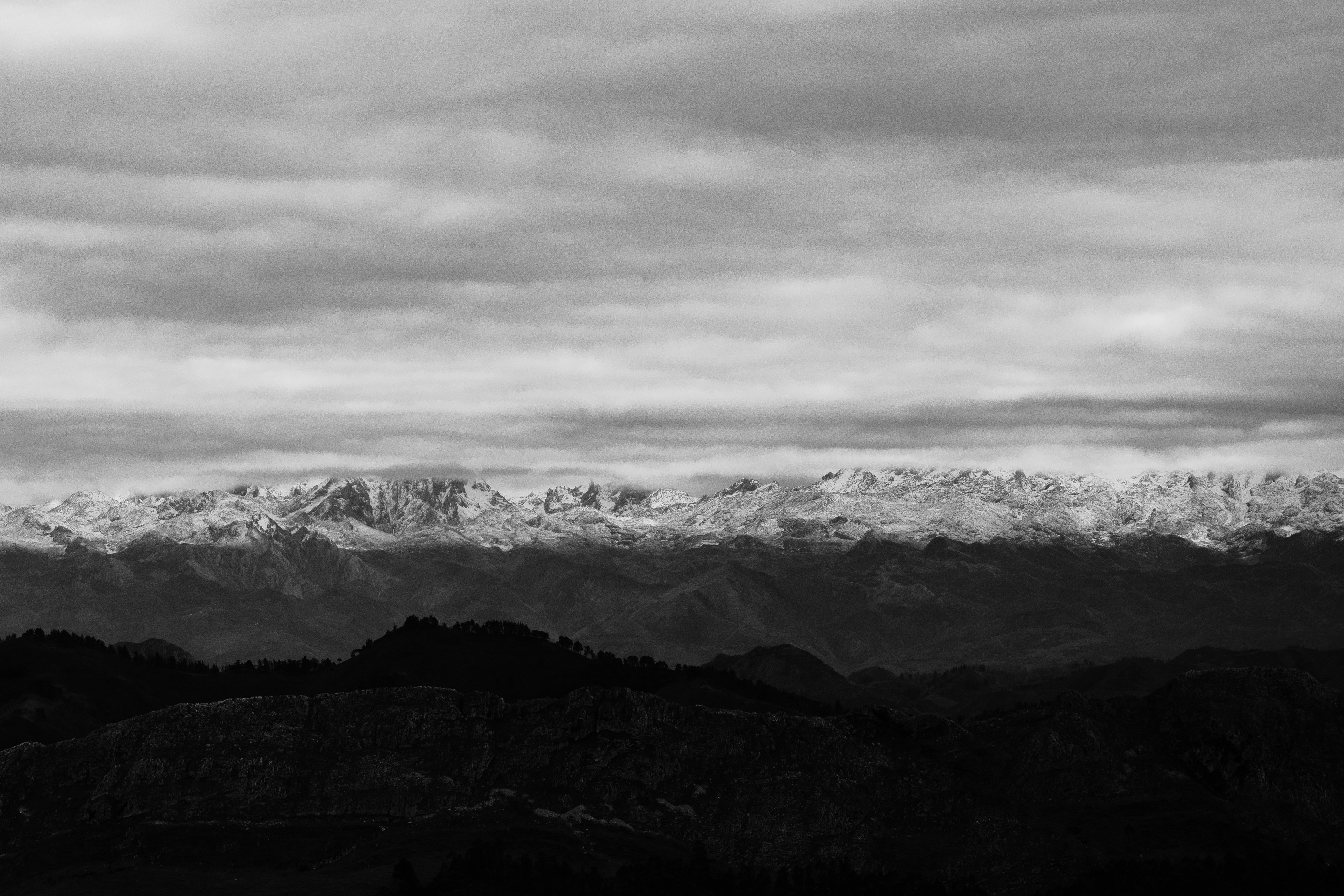 Cordillera nevada bajo un cielo nuboso, con montes oscuros en primer plano, en blanco y negro.