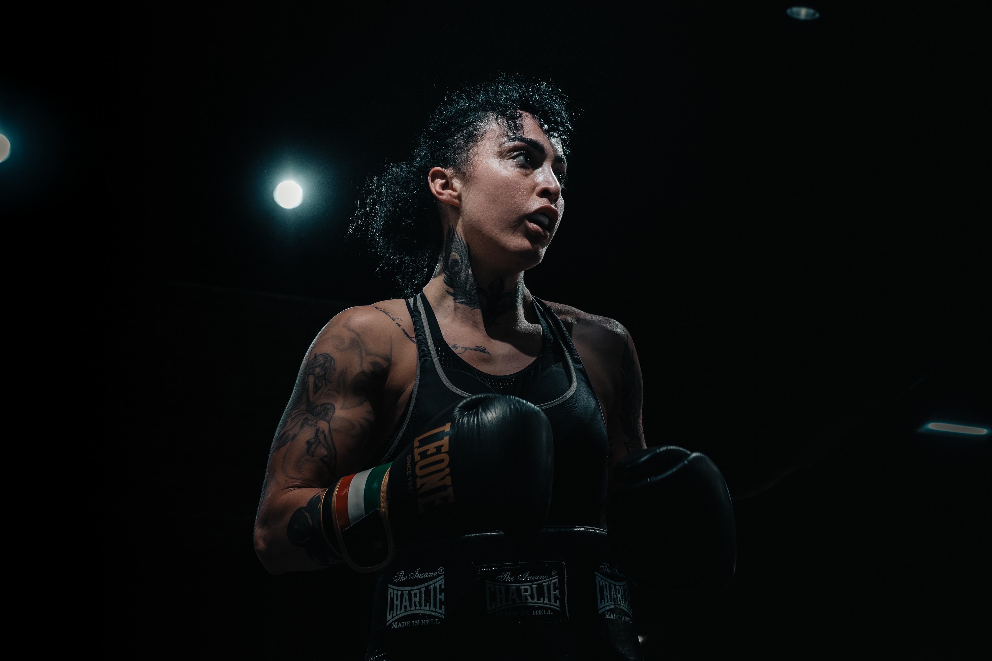 Retrato de una boxeadora con guantes negros y tatuajes bajo las luces del gimnasio.