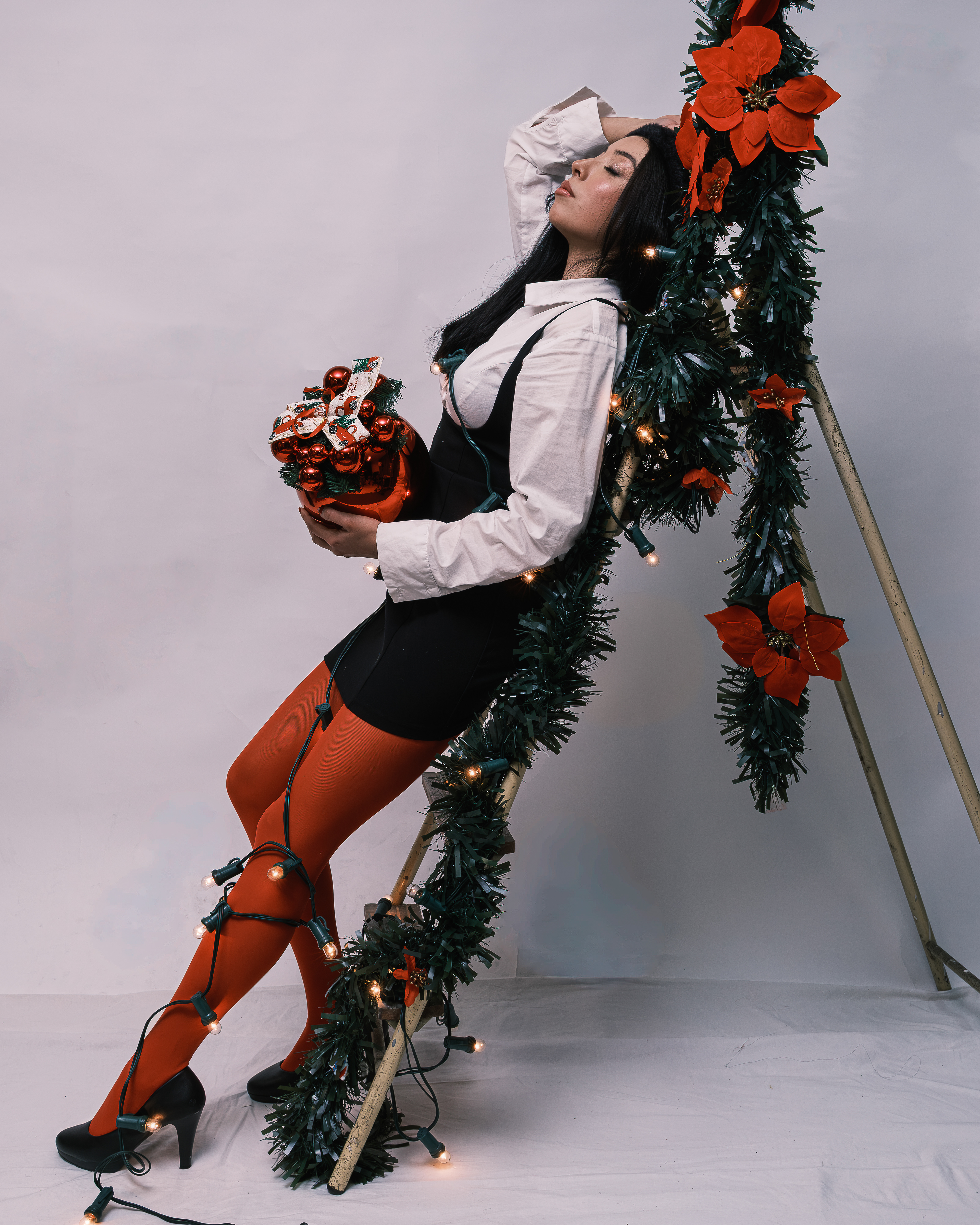 Mujer apoyada en una escalera decorada con guirnaldas y flores rojas, sosteniendo un regalo.
