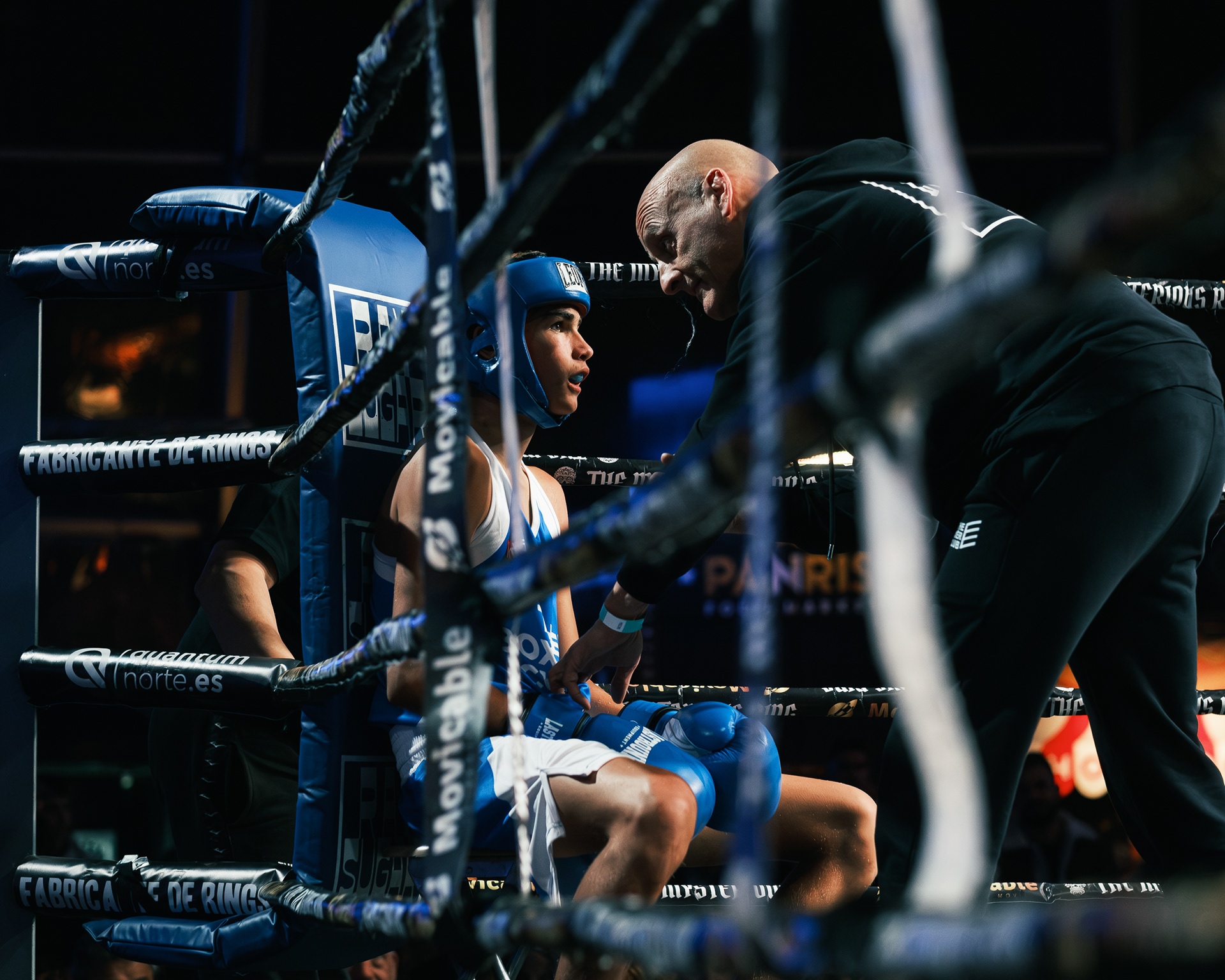 Boxeador vestido de azul sentado en su esquina mientras su entrenador le da indicaciones.
