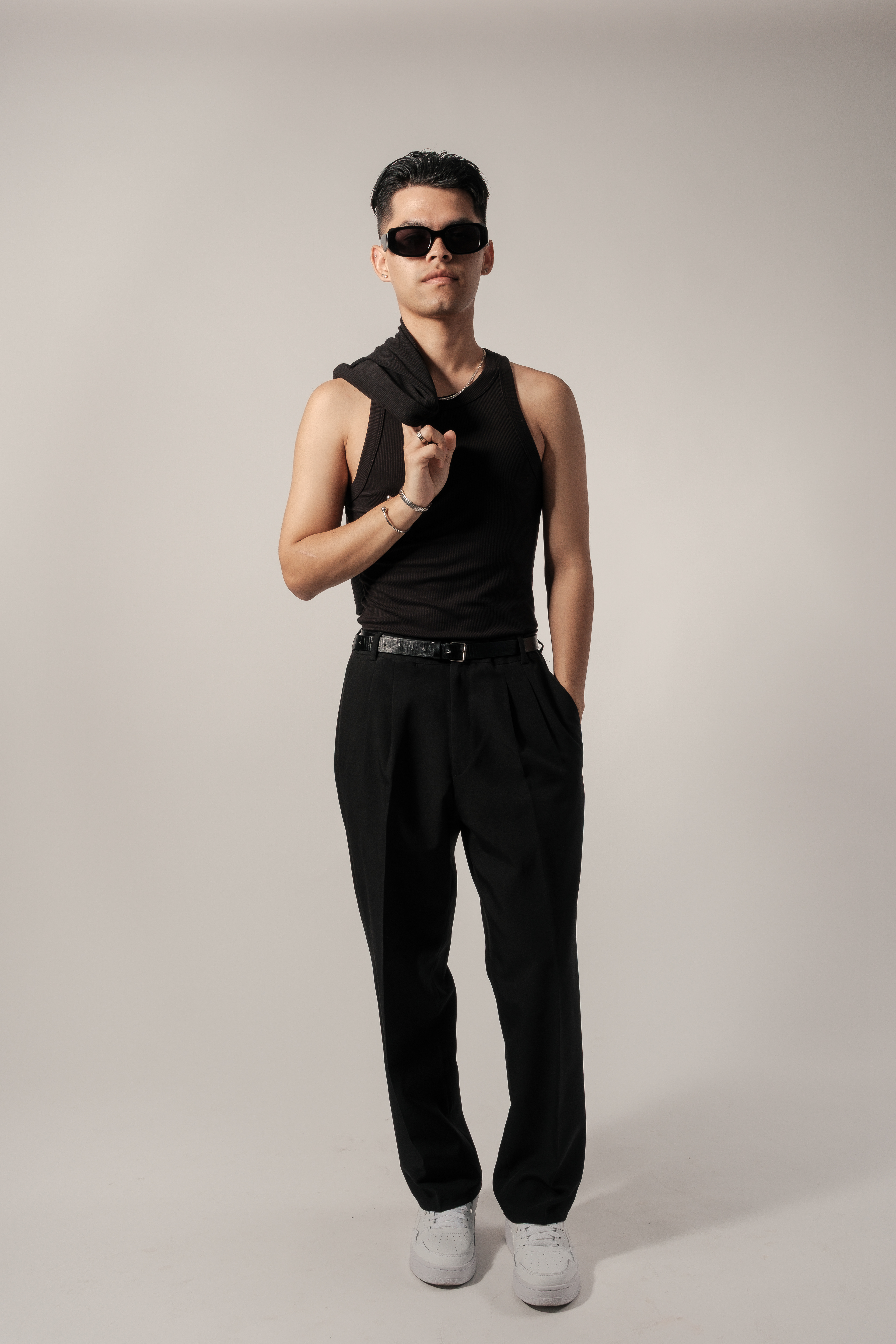 Bryan de pie con camiseta y pantalones negros, gafas de sol y zapatillas blancas, sujetando una prenda al hombro.