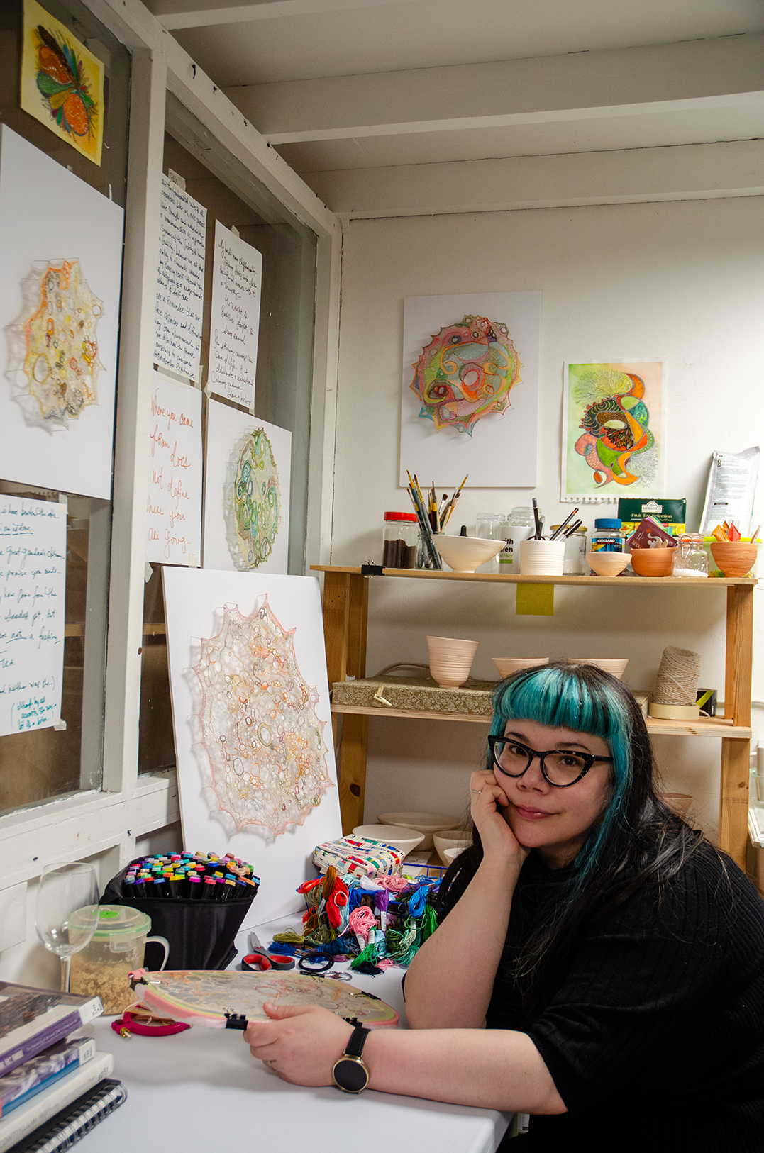 jillian lee adamson sitting in her studio at her desk. Jillian is holding an embroidery hoop and is leaning on her hand which is on the edge of the table. Upon the table is a set of markers, some books, an empty wine glass, a container of oatmeal, and a big pile of embroidery floss. On the walls around the table are embroidery pieces, drawings and handwritten reminders