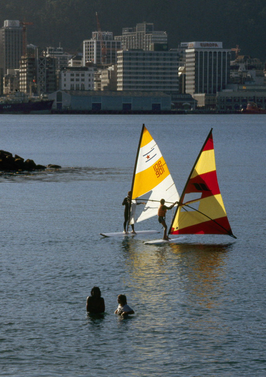 Swimmers and surf sailors off Freyberg Beach, Oriental Vay with Wellington city beyond