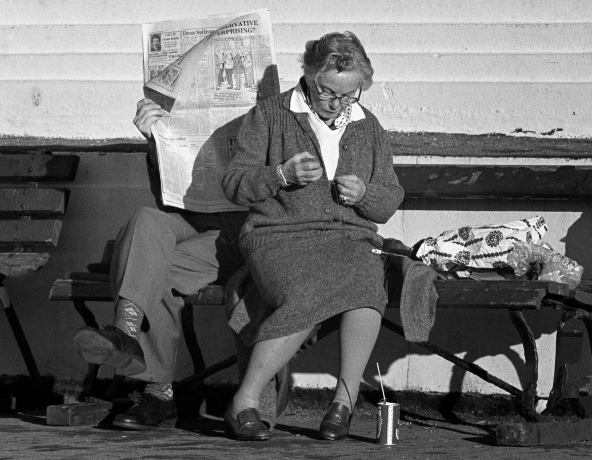 Man reading newspaper, woman knitting, on band rotunda, Oriental Bay, Wellington