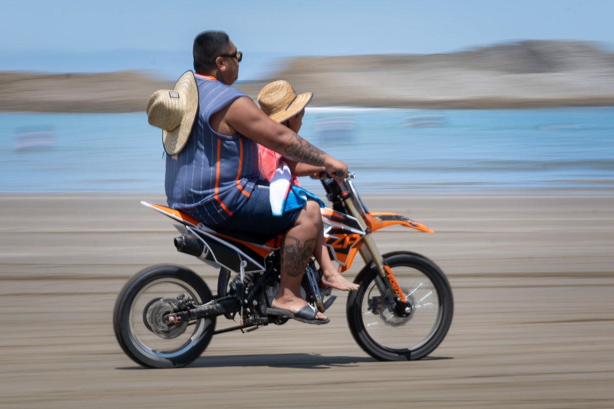 Man with child on a small motorbike on the sand at Castle Point, 2022