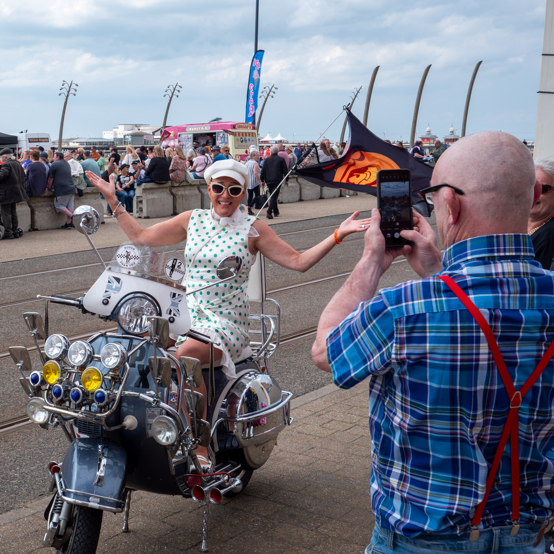 A man in red braces photographs a woman in green spotted dress posing on a classic Mod scooter on the Blackpool Promenade