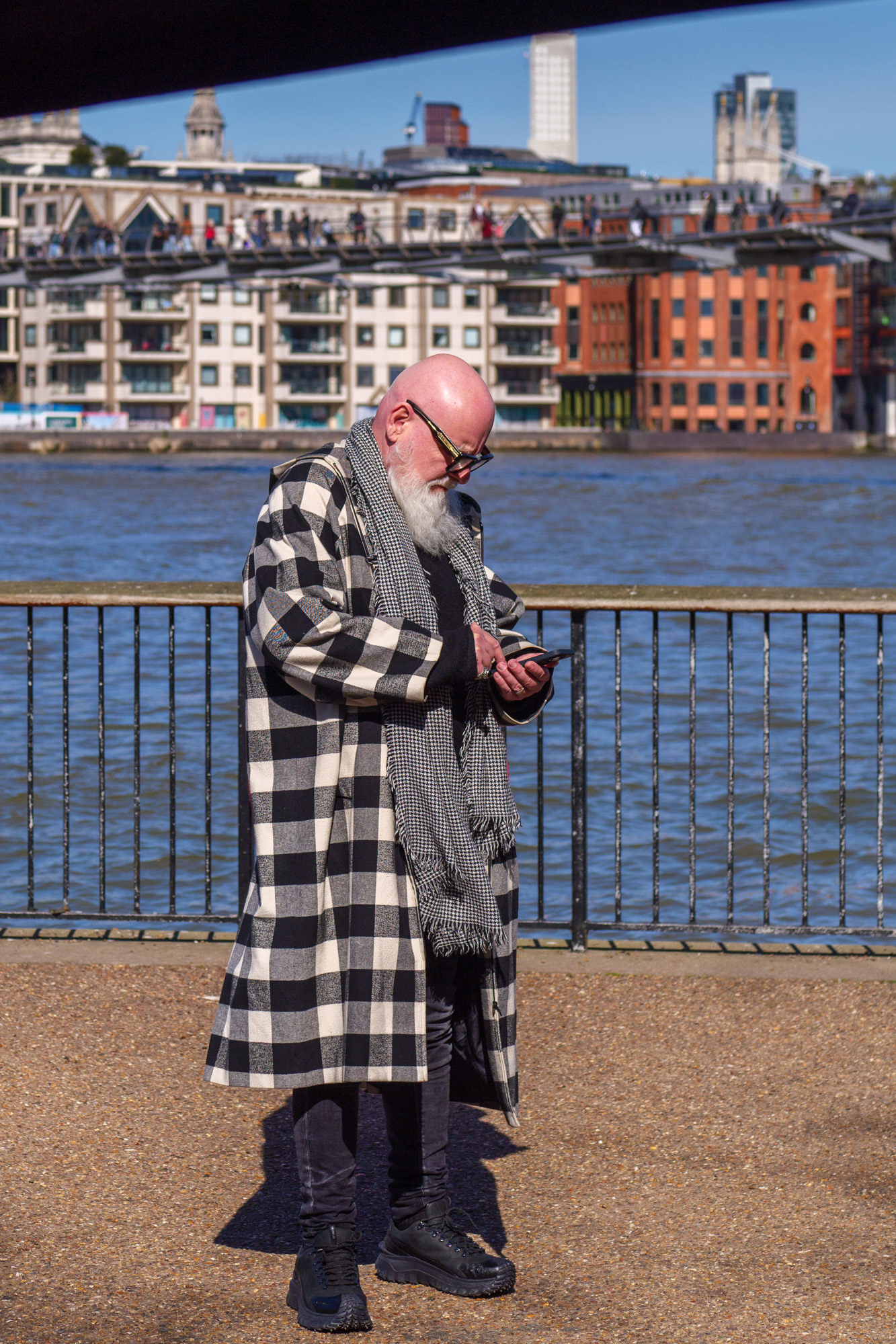 Bald white man with white beard and check coat looking at his phone beside the Thames in London
