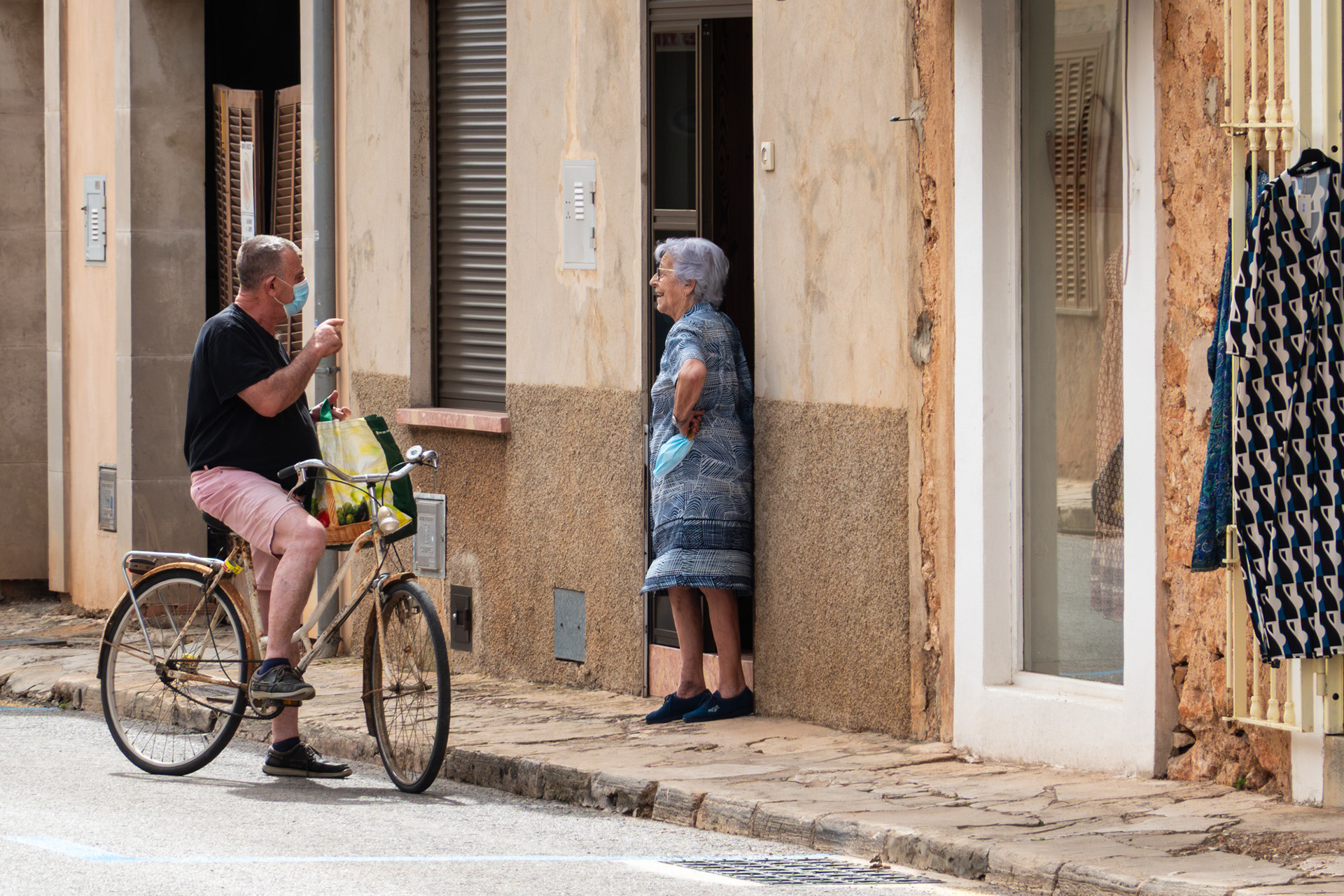 Man in pink shorts and a COVID mask on bicycle chatting to older woman in a doorway of house in Campos, Mallorca