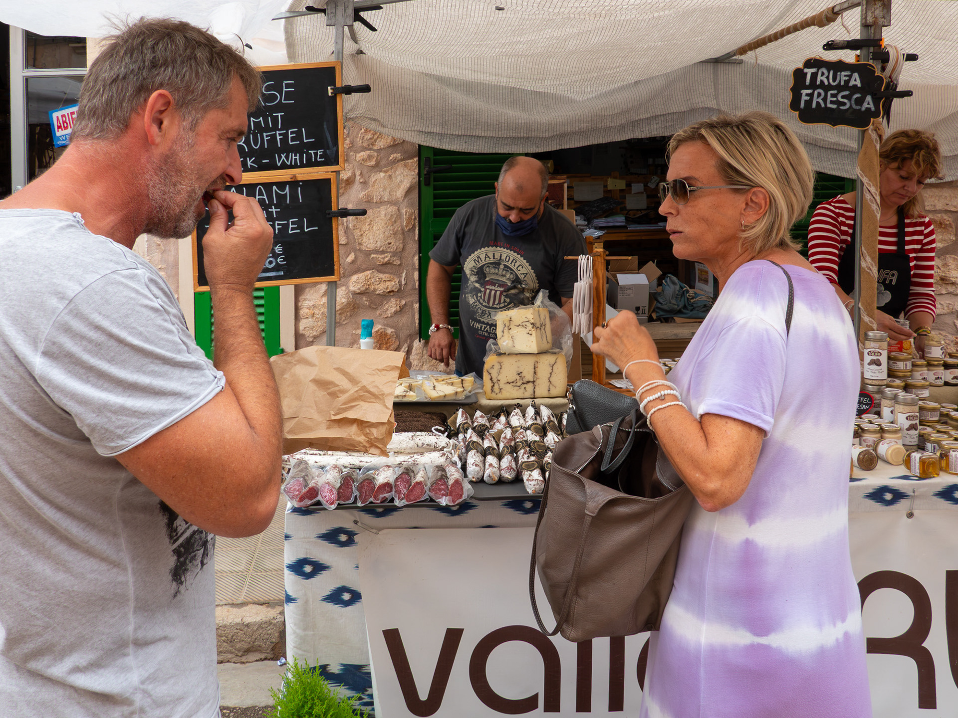 Man tasting food at a street delicatessen in Santanyi, Mallorca while his wife with purse waits for his opinion