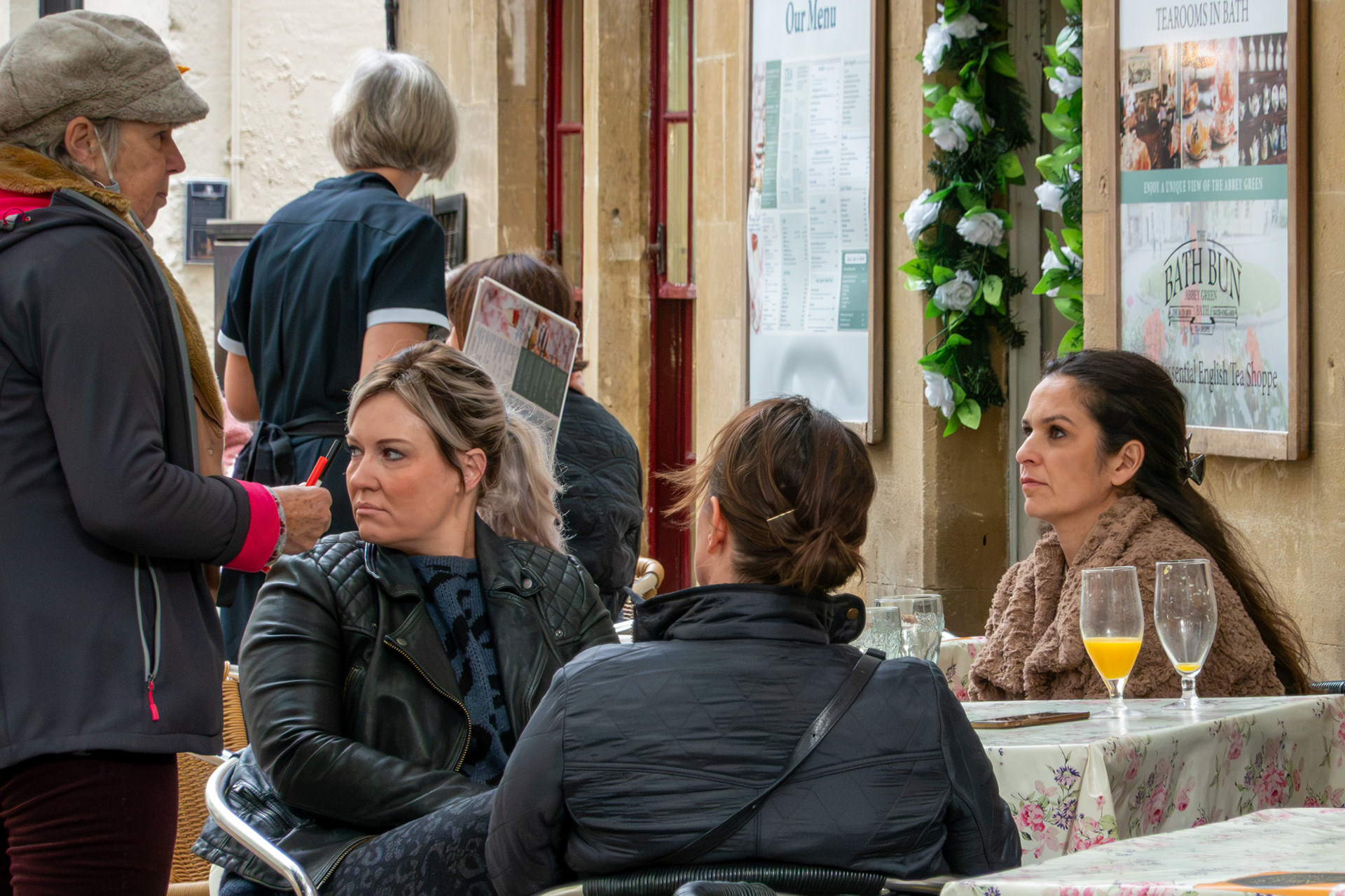 In Bath, a seated customer and a standing woman in winter coats lock eyes outside a tearoom while a friend looks away