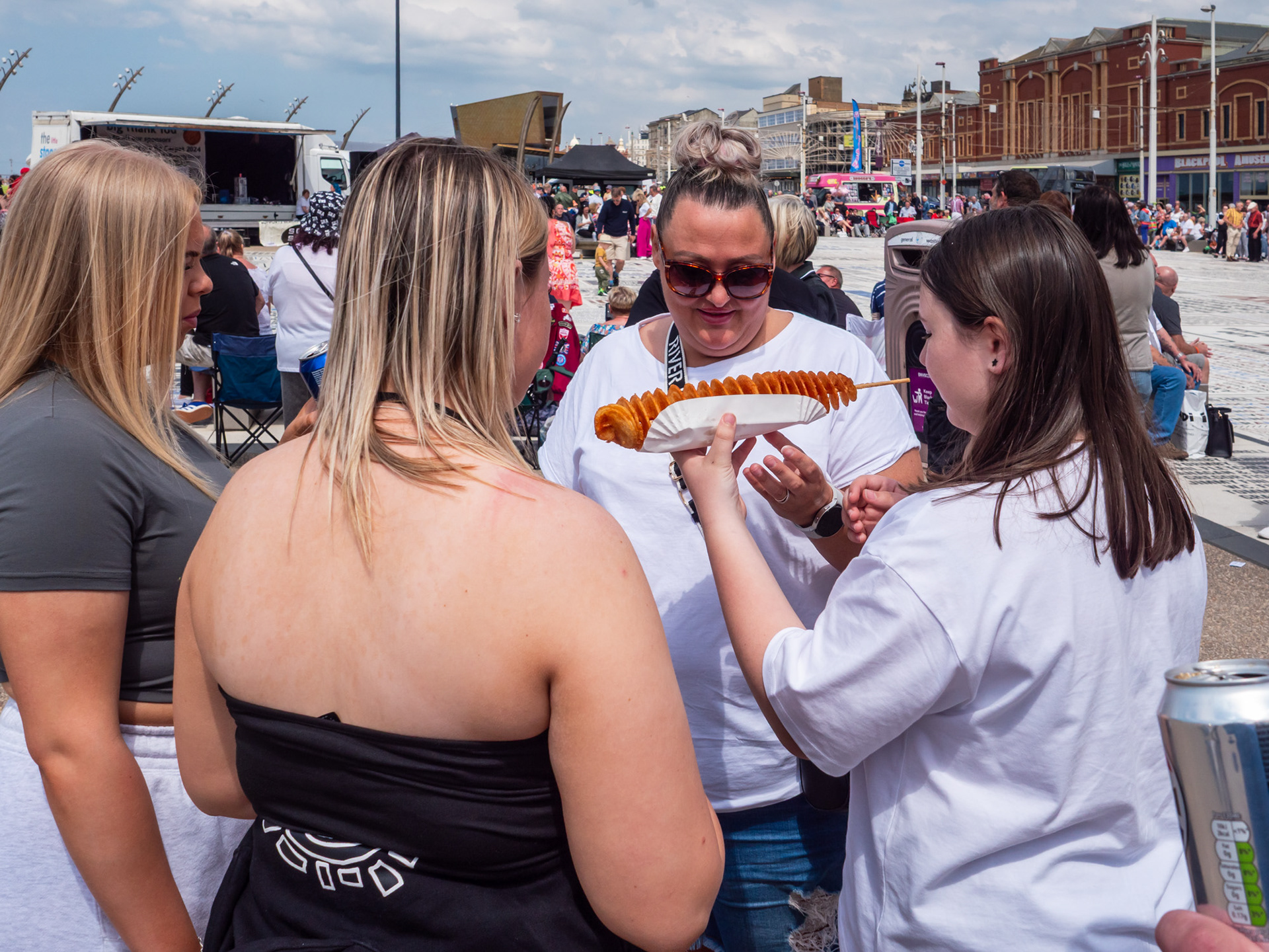 Four women standing by the busy Comedy Carpet in sunny Blackpool, sharing and admiring a tornado potato