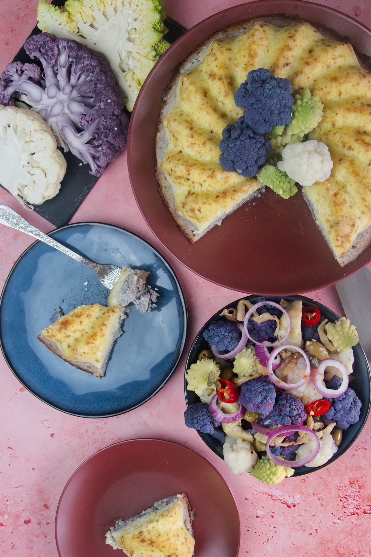 Overhead view of a savory cauliflower sformato, sliced portions on small plates, and a colorful mixed cauliflower salad with red onions and olives on a pink surface.