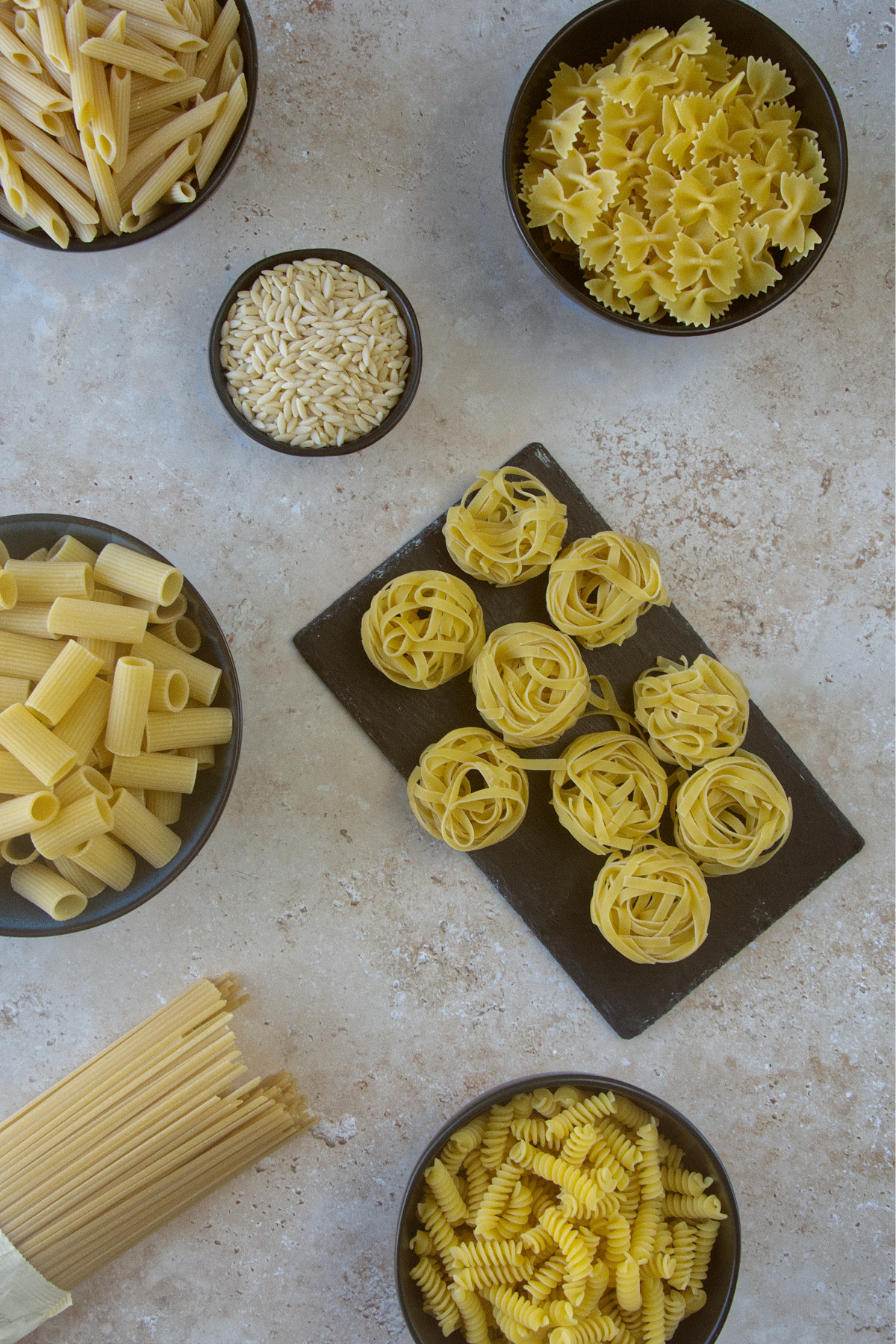 A variety of dry pasta shapes, including bowls of penne, farfalle, rigatoni, fusilli, and orzo, alongside bundles of spaghetti and nests of tagliatelle.