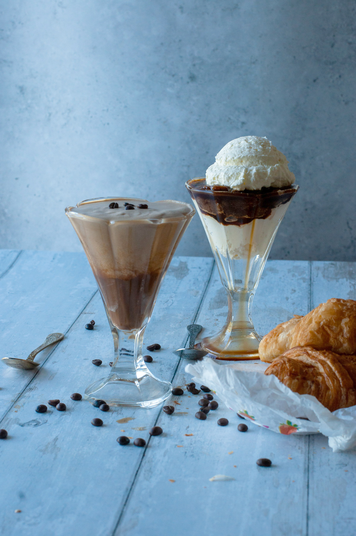 Coffee cream and layered coffee granita with whipped cream in glass cups on a light blue wooden table, surrounded by scattered coffee beans and croissants.
