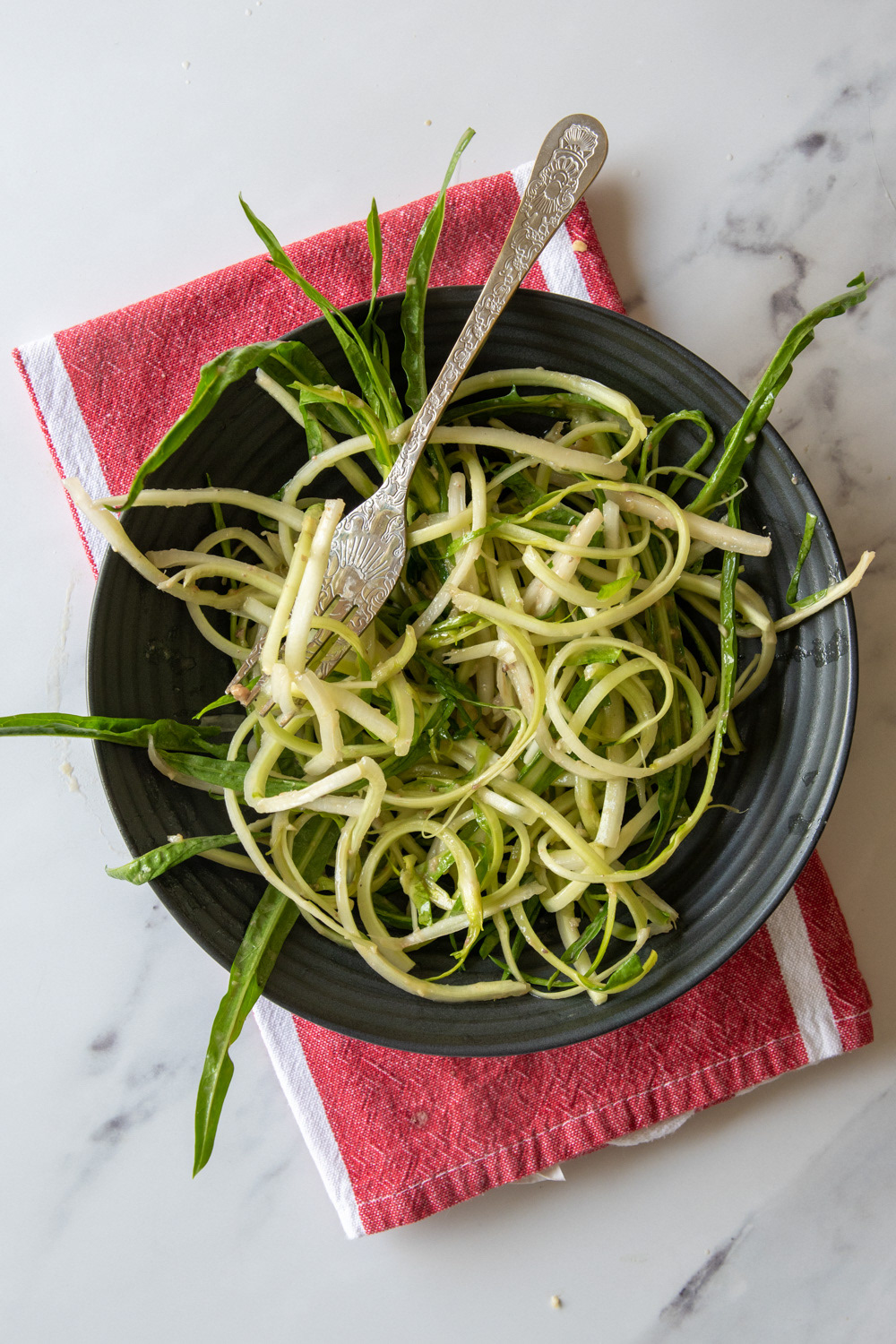 Overhead view of curled puntarelle salad in a black bowl with a silver fork, resting on a red and white striped napkin over a white marble surface.