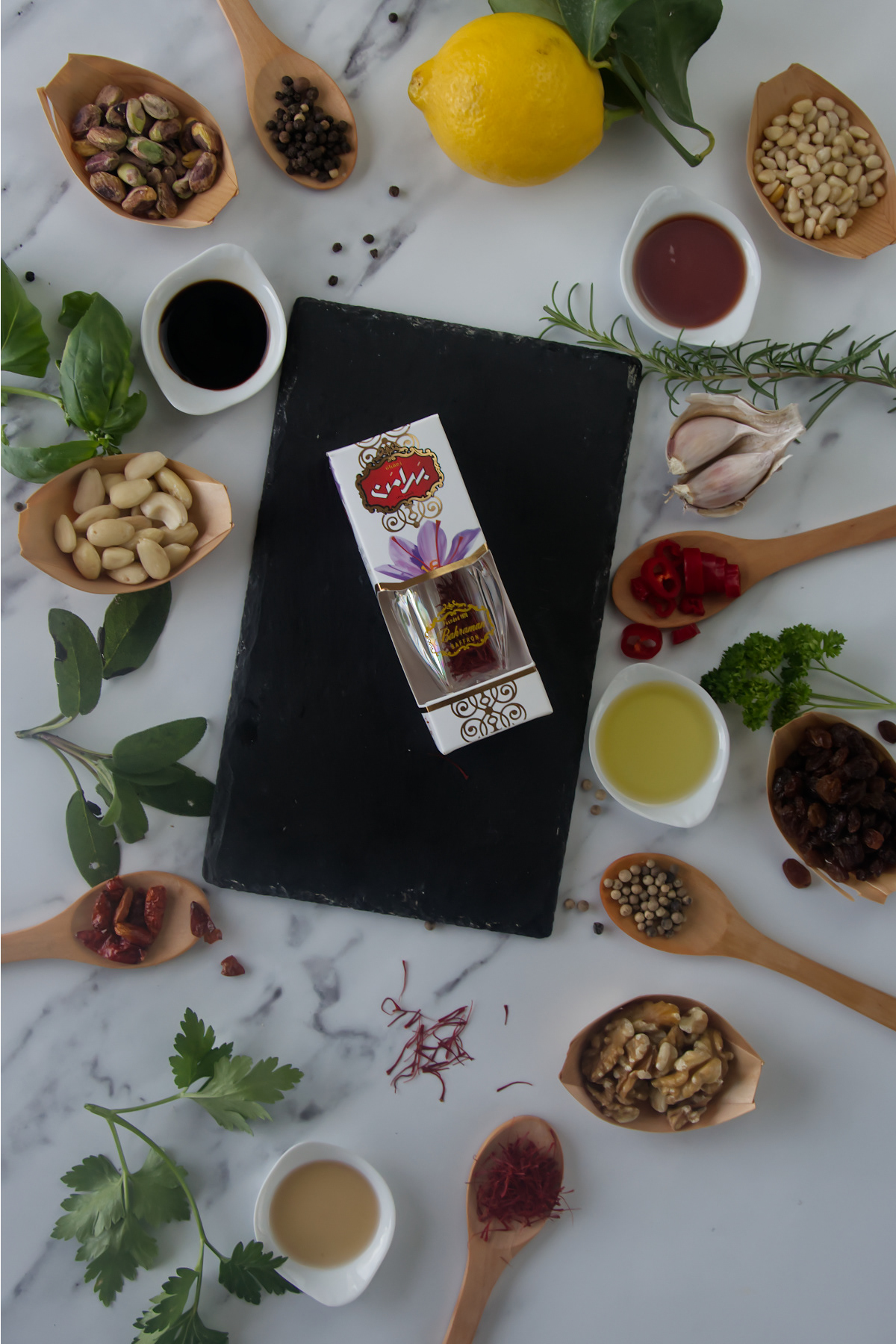 A box of saffron on a black slate surrounded by wooden spoons of spices, nuts, and dried fruit, alongside fresh herbs, a lemon, and small bowls of oil and vinegar on a marble surface.