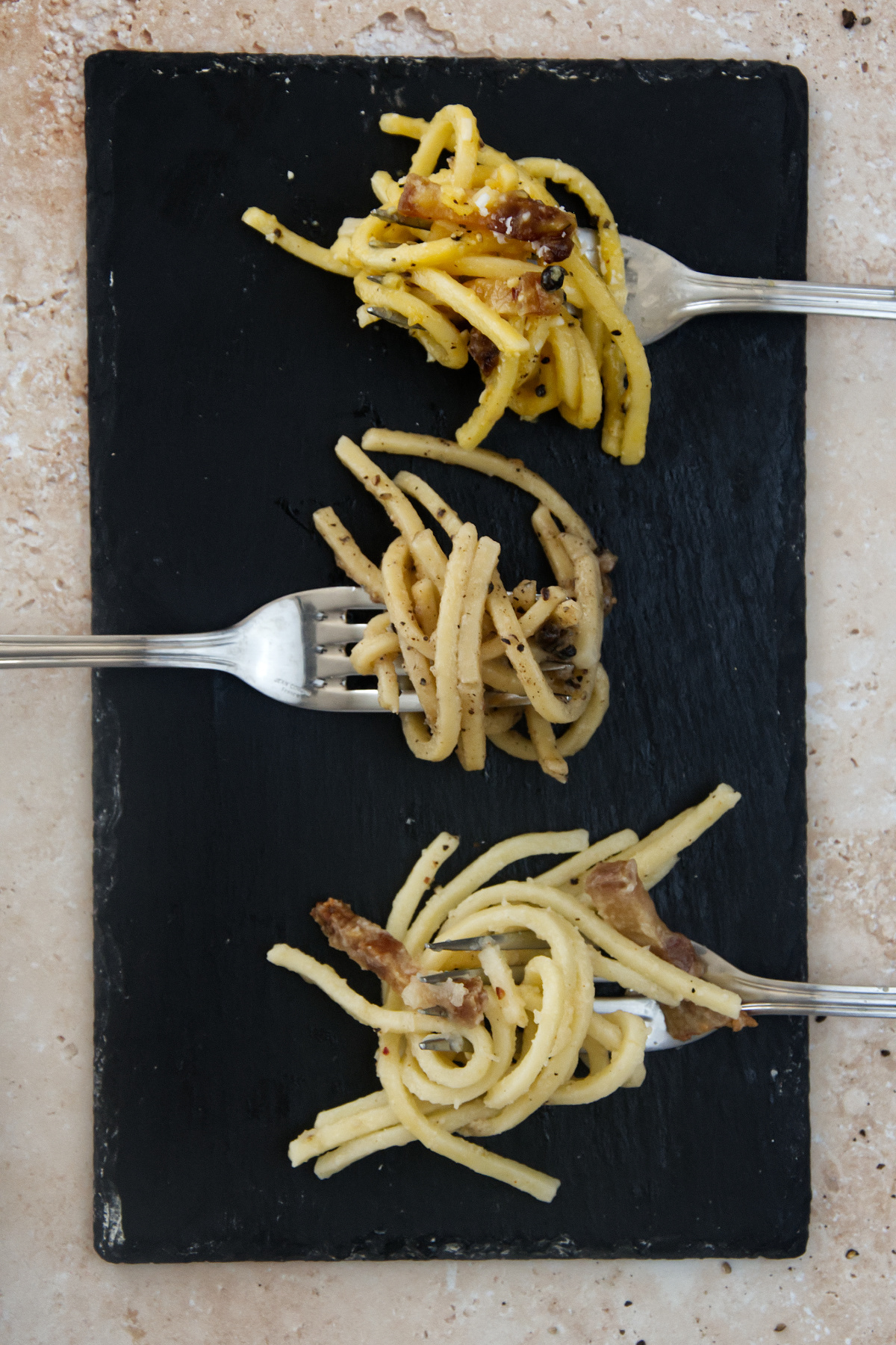 Top-down view of three silver forks arranged vertically on a black slate board, each twirled with a distinct Roman pasta dish: Carbonara, Cacio e Pepe, and Gricia. The board rests on a light, textured stone background.