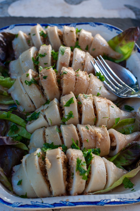 Close-up view of sliced, stuffed calamari arranged on a bed of mixed salad greens in a rectangular white serving dish with a blue patterned edge. The dish is garnished with fresh chopped parsley, and a silver serving fork rests on the right side.
