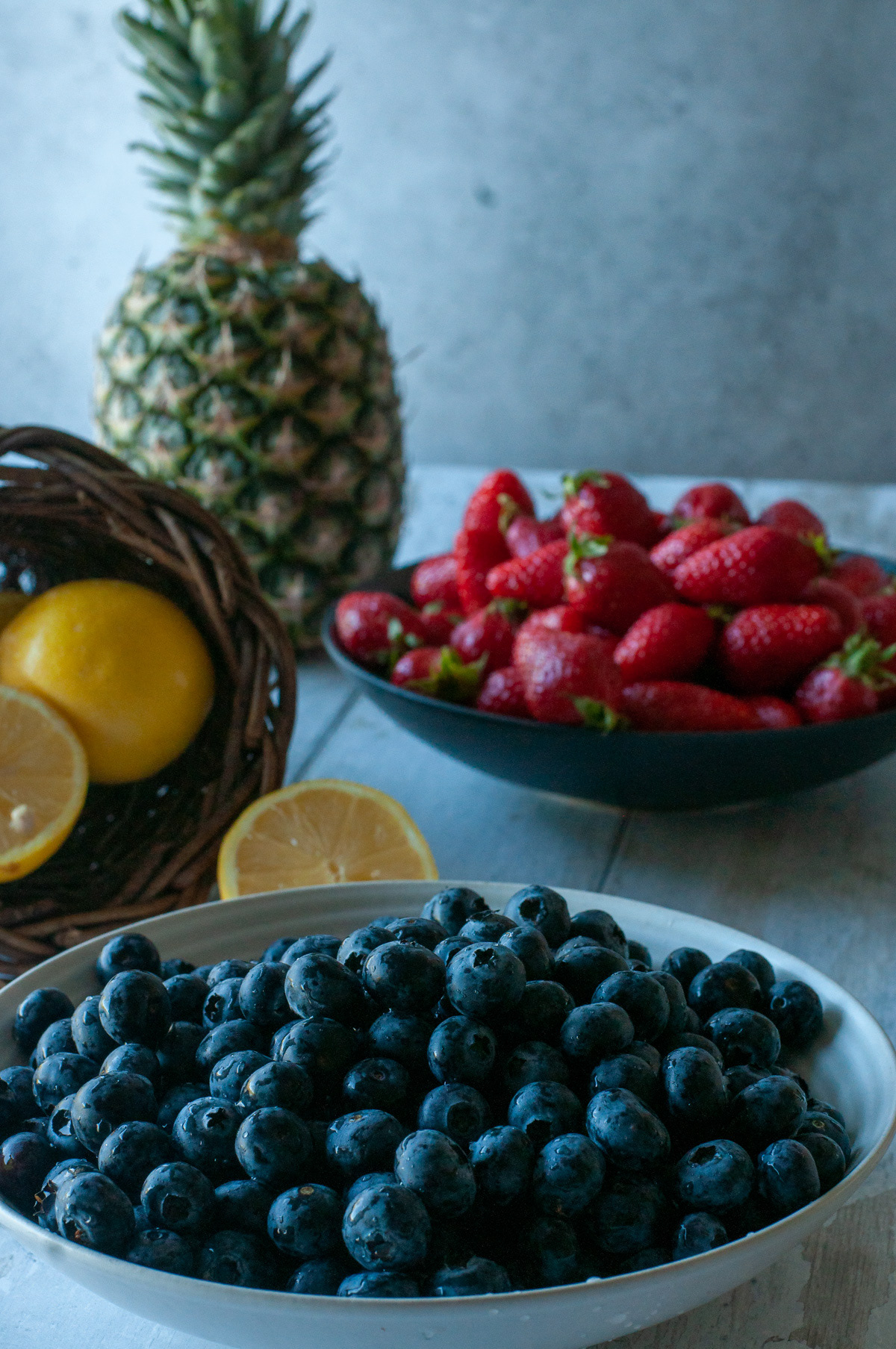 A bowl of fresh blueberries in the foreground, with strawberries, a pineapple, and lemons in a basket in the background.