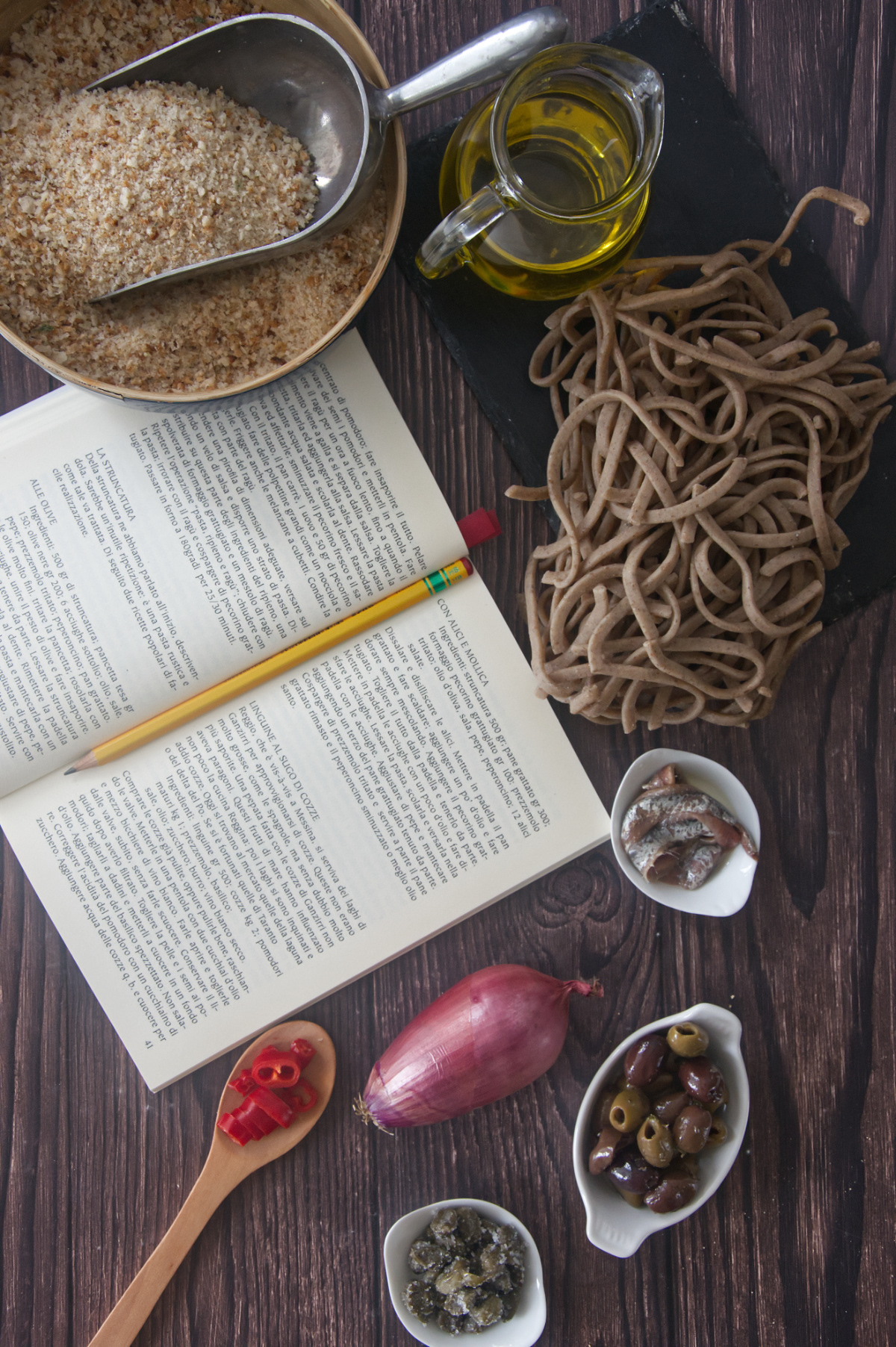 Top-down view of pasta ingredients including dry noodles, olives, onions, and an open cookbook.