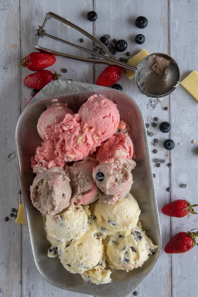 Three flavors of gelato in a metal pan, surrounded by fresh berries, chocolate chips, and an ice cream scoop on a white wooden surface.