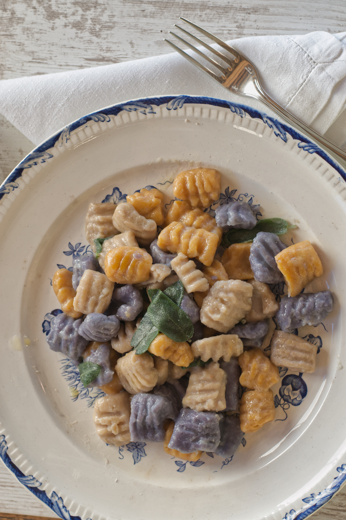 Overhead view of a portion of homemade gnocchi made from blue potatoes, squash, and chestnuts, garnished with fresh sage leaves. The pasta is served on a white plate with a blue patterned rim, accompanied by a silver fork and white cloth napkin on a light wooden surface.