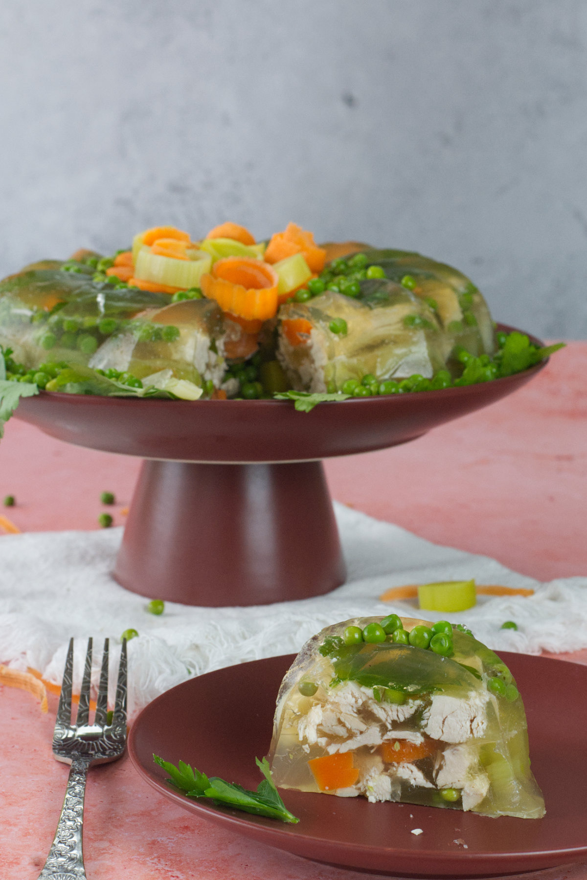 Sliced chicken aspic with peas and carrots on a dark red pedestal, with a single plated portion and silver fork in the foreground on a pink surface.