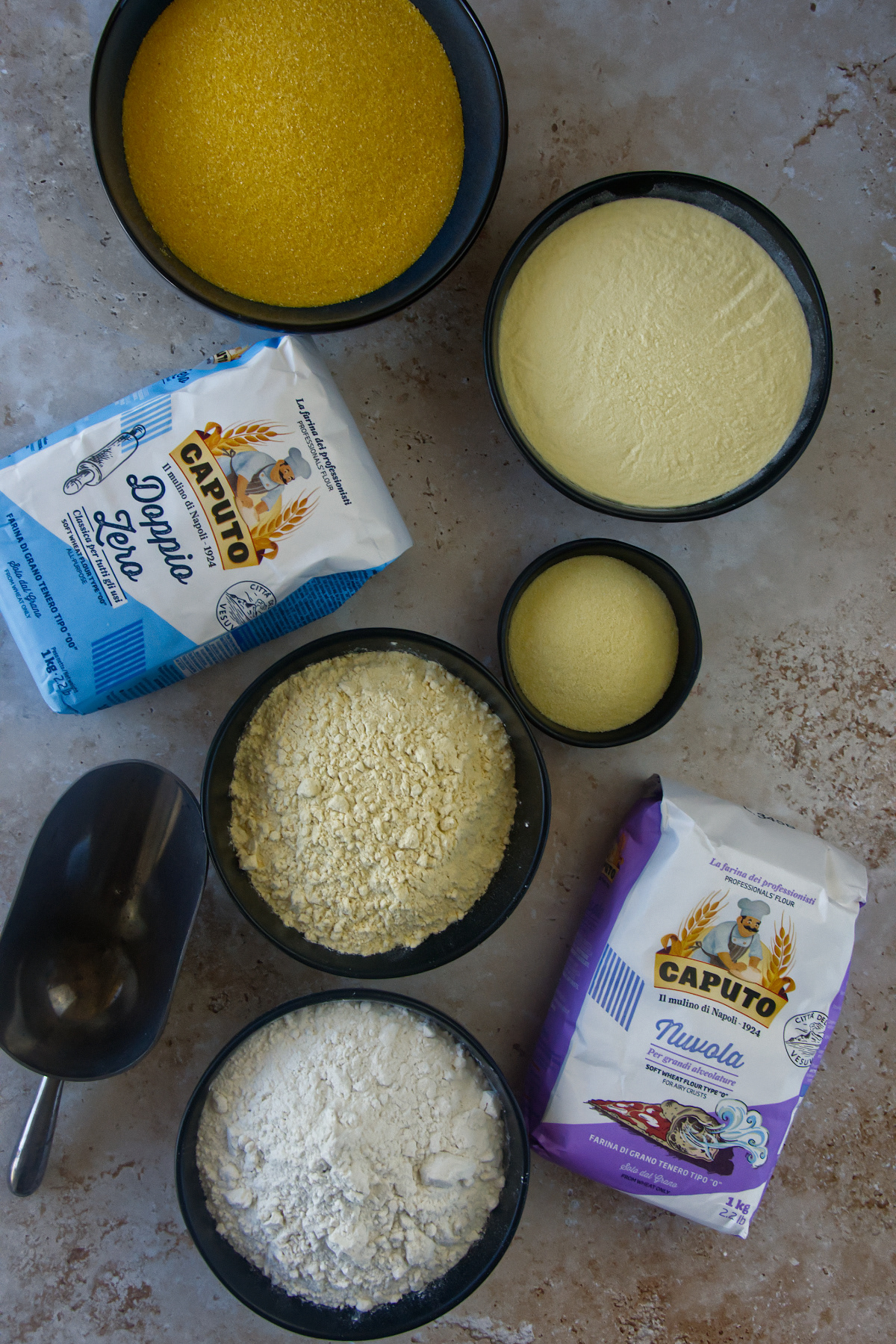 Top-down flat lay of various types of flour and yellow cornmeal in black bowls, accompanied by bags of Caputo Italian flour and a metal scoop on a stone countertop.