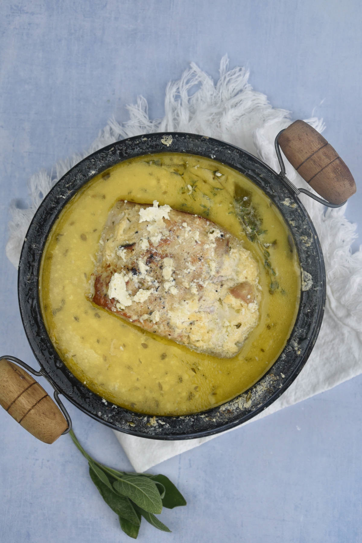 Overhead view of pork cooked in milk sauce with herbs in a black pot with wooden handles, resting on a white cloth and light blue surface.