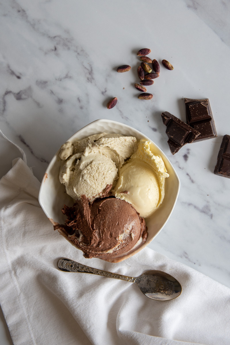 Overhead view of chocolate, vanilla, and pistachio gelato in a white bowl on a marble surface, surrounded by chocolate chunks, shelled pistachios, and a silver spoon.
