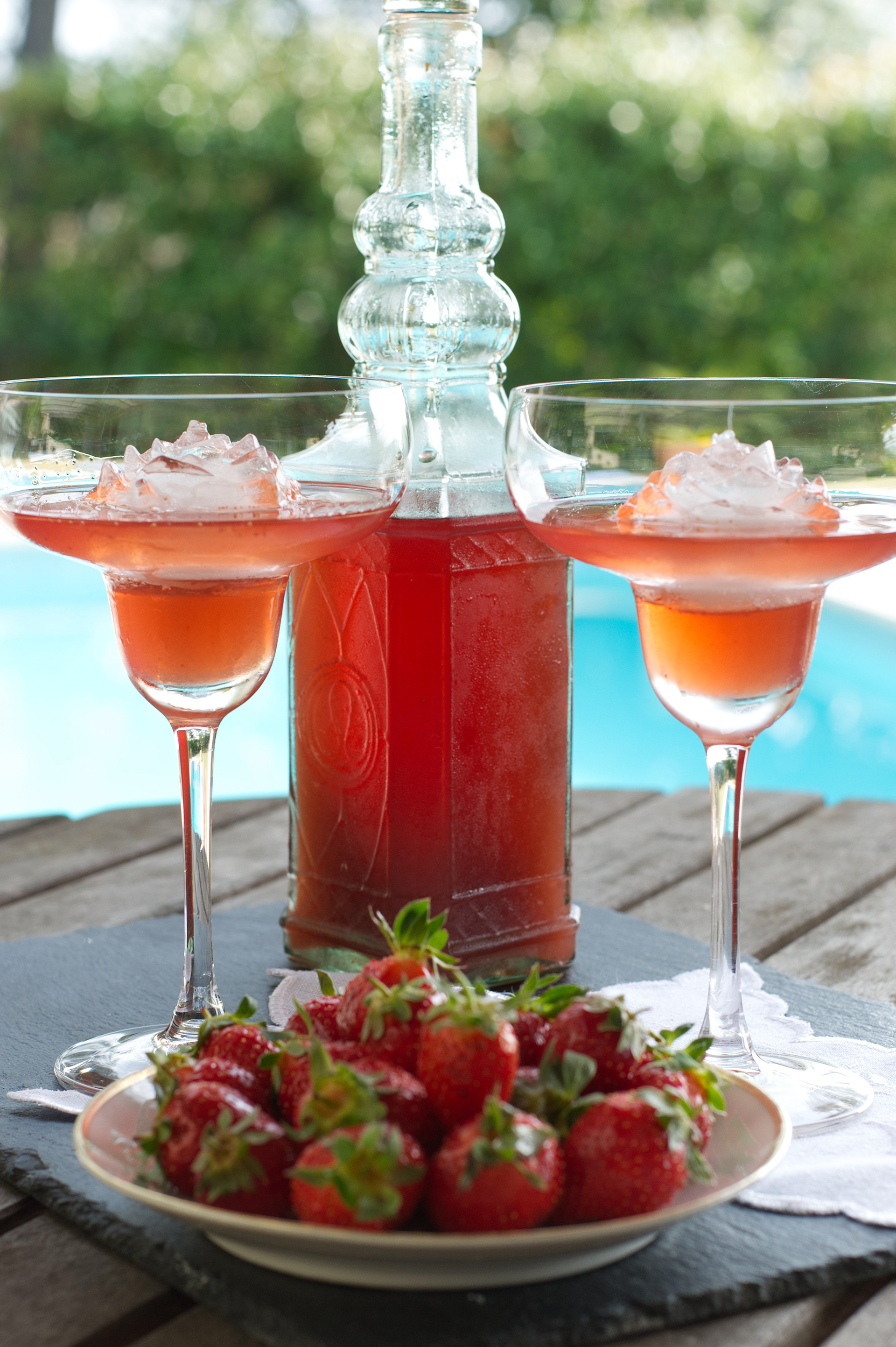 Chilled strawberry liqueur served in stemmed glasses with fresh strawberries on a terrace overlooking a pool.