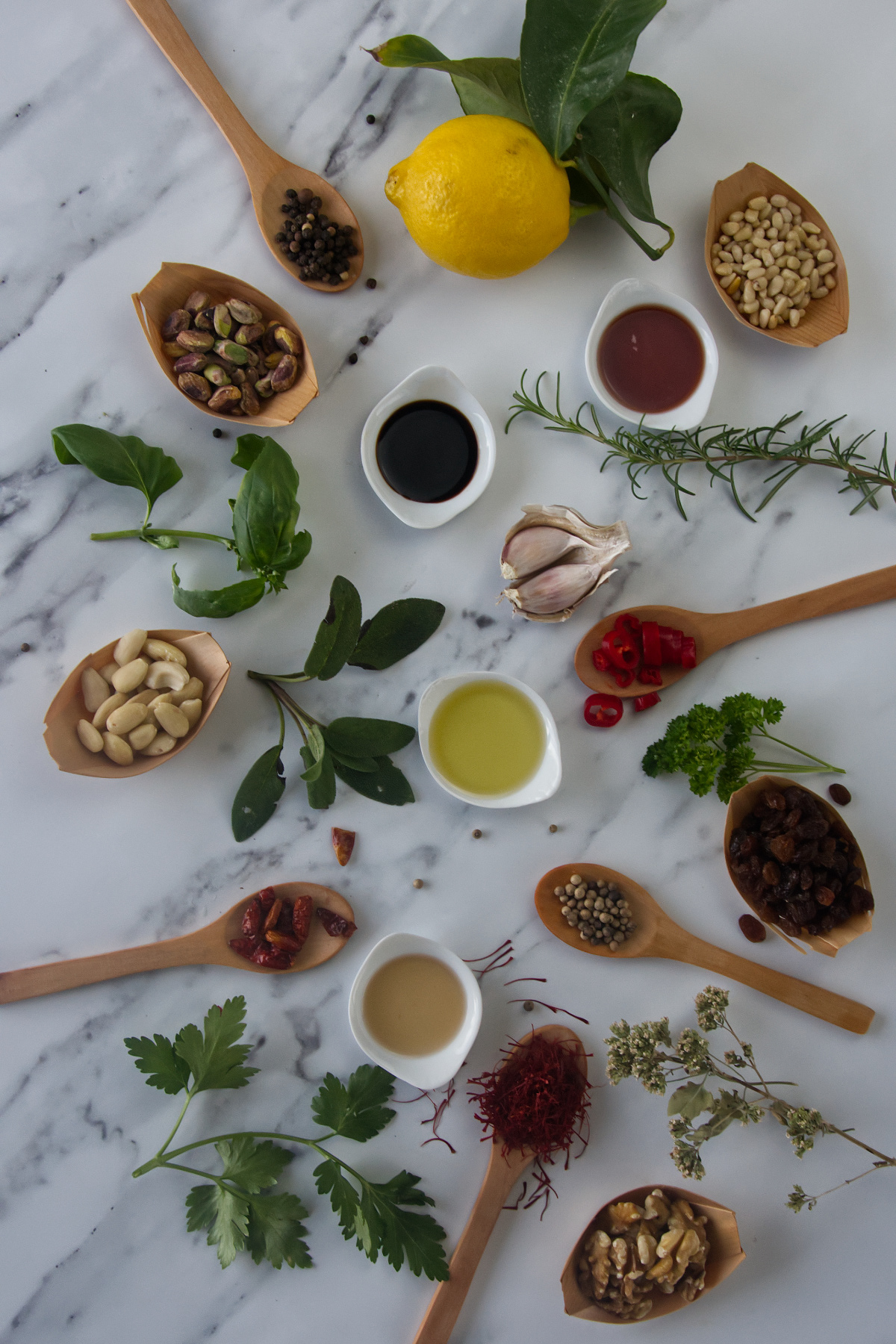 Overhead view of a lemon, garlic, and oils on marble, surrounded by spoons and bowls containing pine nuts, pistachios, almonds, walnuts, raisins, saffron, peppercorns, chili, and fresh herbs.
