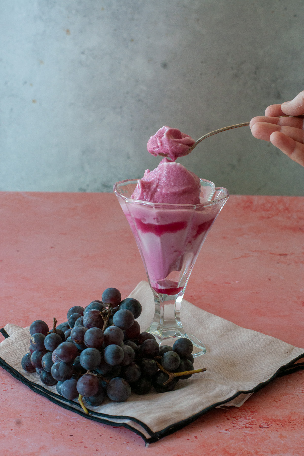 A hand lifting a spoonful of purple grape sorbet from a tall glass dish, resting next to a bunch of fresh dark grapes on a pink surface.