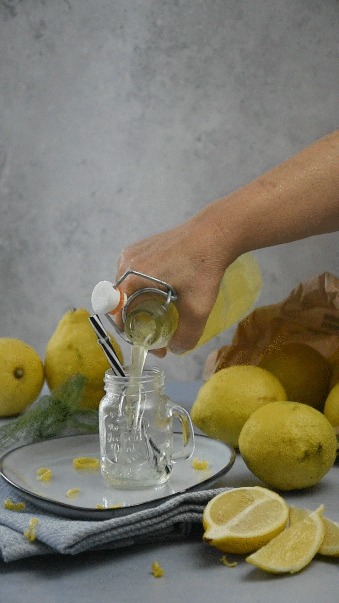 A hand pouring chilled limoncello from a flip-top bottle into a small glass jar with a straw, surrounded by fresh whole and sliced lemons.