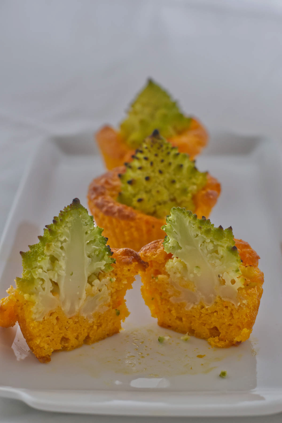 Three savory carrot muffins with Romanesco broccoli florets on a white rectangular plate, with the front muffin cut in half.
