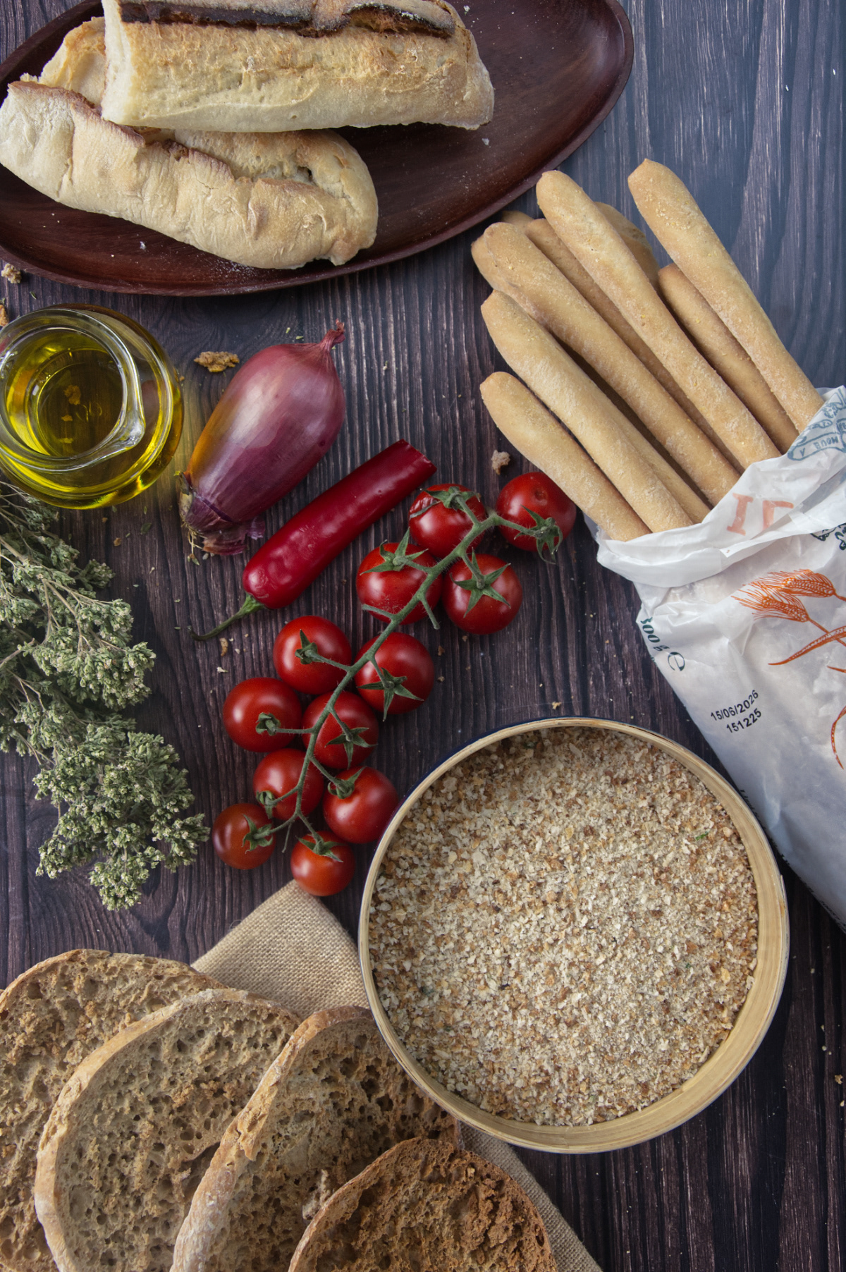 A rustic wooden table set with staple ingredients including bread slices, a bowl of breadcrumbs, grissini, cherry tomatoes on the vine, a red onion, chili pepper, olive oil, and dried herbs.