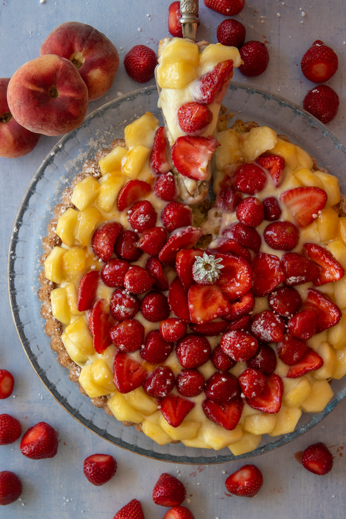 Overhead view of a sliced fruit tart topped with cream, strawberries, and peaches in a glass dish, surrounded by fresh fruit.
