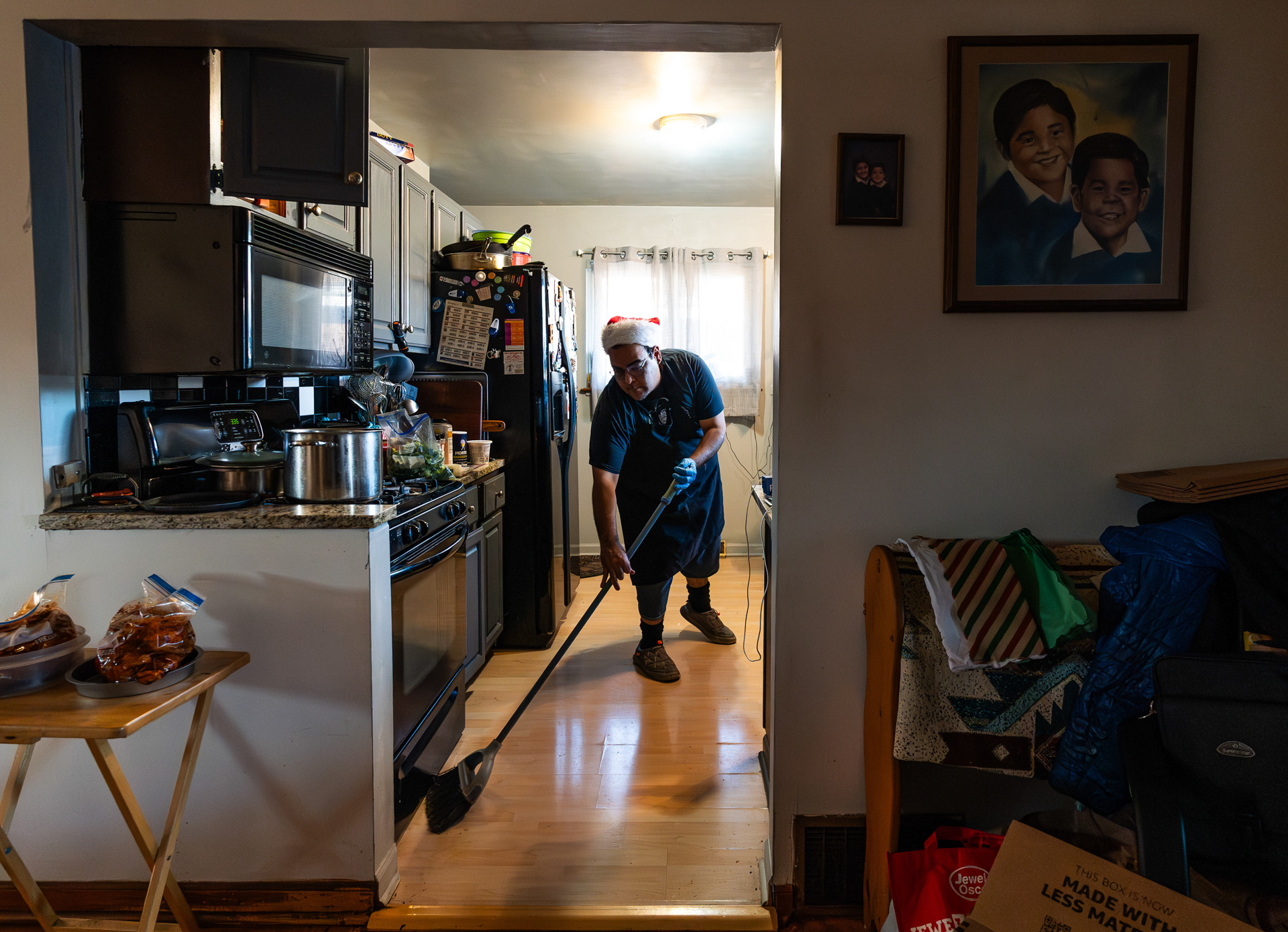 Daniel Cano sweeps the kitchen floor at his home in Westchester, Illinois on November 17, 2023. A photo and a drawing of Cano and his brother as children hang on the wall.