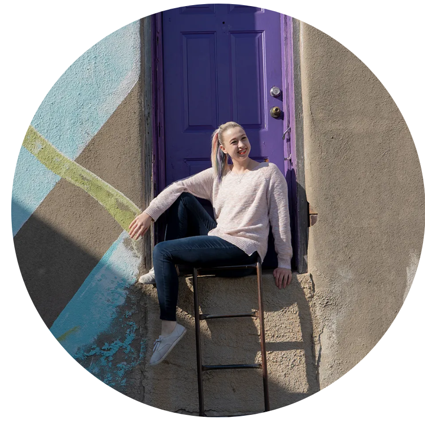 Foreground features Bailey Vaughan in pink sweater with jeans and white tennis shoes. Her hair is pulled back in a pony tail. Background features a cement wall with a purple door half way up. Bailey sits on the ledge of the door.
