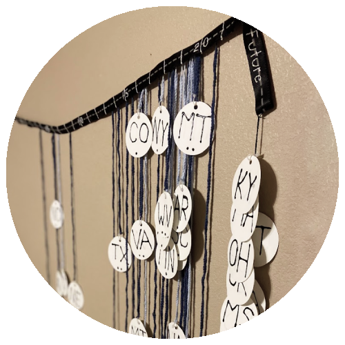Foreground features a dog leash with embroidered numbers representing a timeline with crochet chains dangling down. On the end of each chain is a paper dog tag with a US state abbreviation. Background features a tan wall..