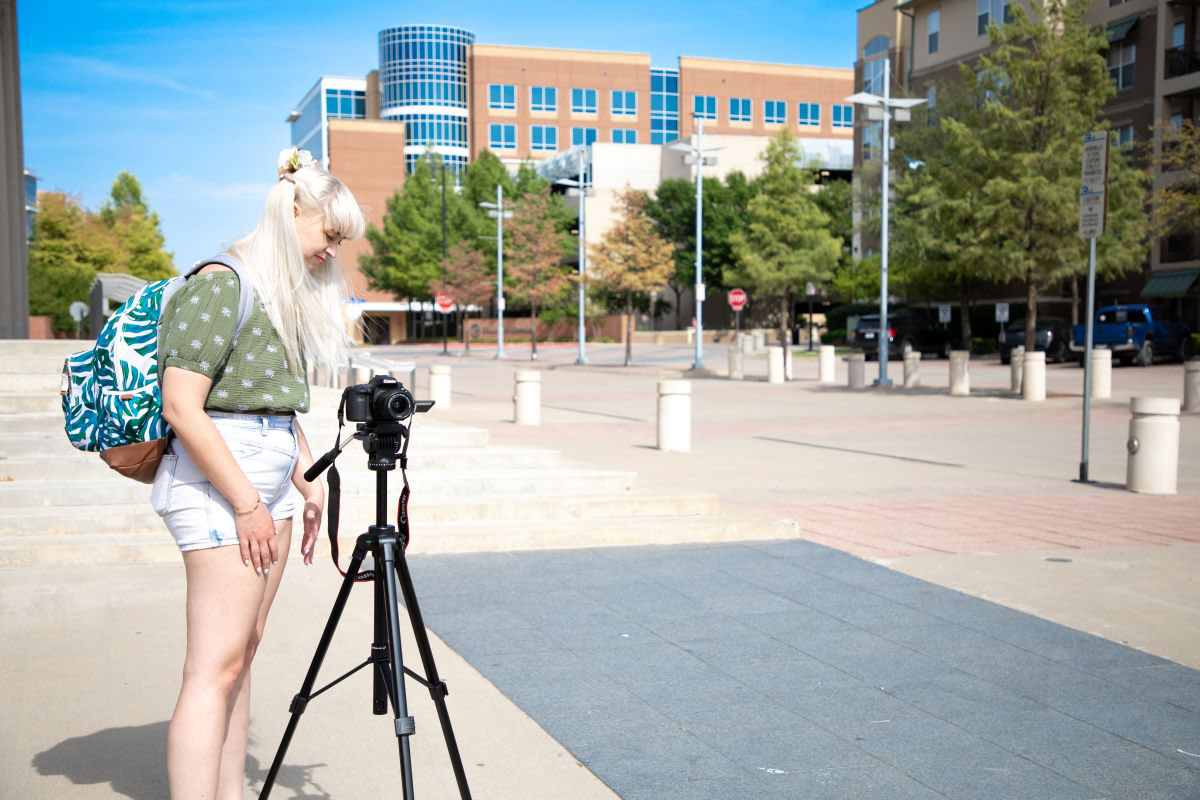 Foreground features a young lady with bangs and blonde hair standing at a DSLR camera on a tripod. Background features a town square with sidewalks and lined with small trees.