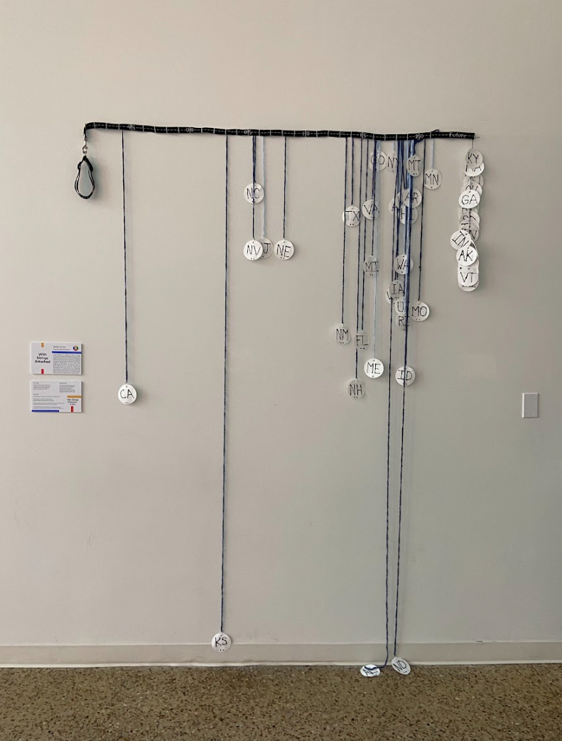 Horizontal dog leash with embroidery hangs on a white wall. Crochet chains of various lengths with paper dog tags hand from the leash. Next to the sculpture is a plaque on the wall displaying the artwork's title and description.