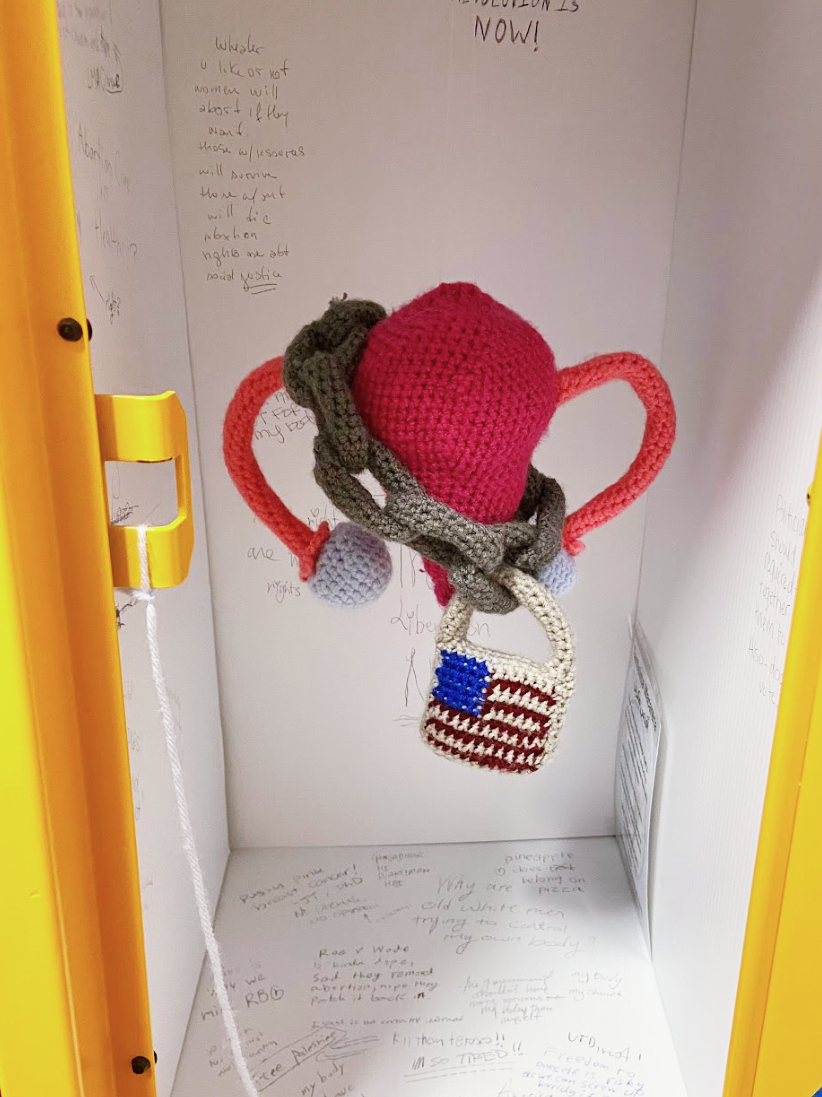 Inside a yellow locker is a pink crochet uterus and gray chain featuring a lock with the US flag. In the locker is a pen inviting people to write on the white panels that line the inside of the yellow locker. The walls feature various hand written messages about participant's view on reproductive rights in the US.