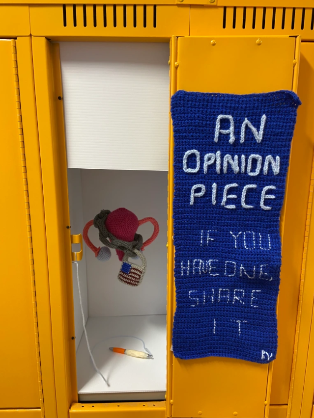 Yellow locker door displays a dark blue crochet banner with the words "An Opinion Piece. If you have one share it." written in light blue crochet yarn. Inside the yellow locker are white panels with a crochet uterus and chain featuring a lock with the US flag. In the locker is a pen inviting people to write on the white panels.