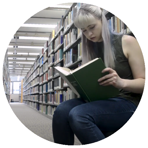 Foreground features young lady with bangs and blonde hair reading a book wth a dark green cover. She sits in a library surrounded by books.