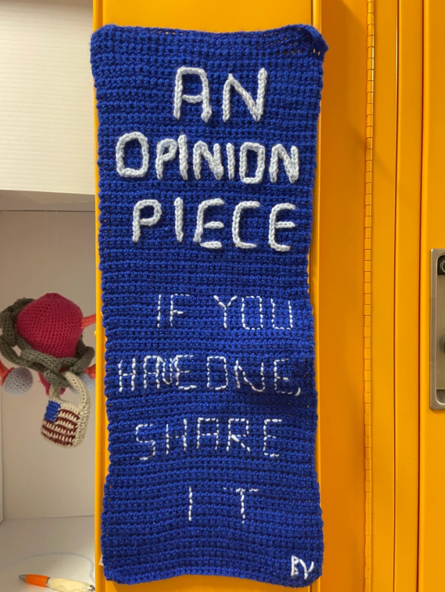 Yellow locker door displays a dark blue crochet banner with the words "An Opinion Piece. If you have one share it." written in light blue crochet yarn.