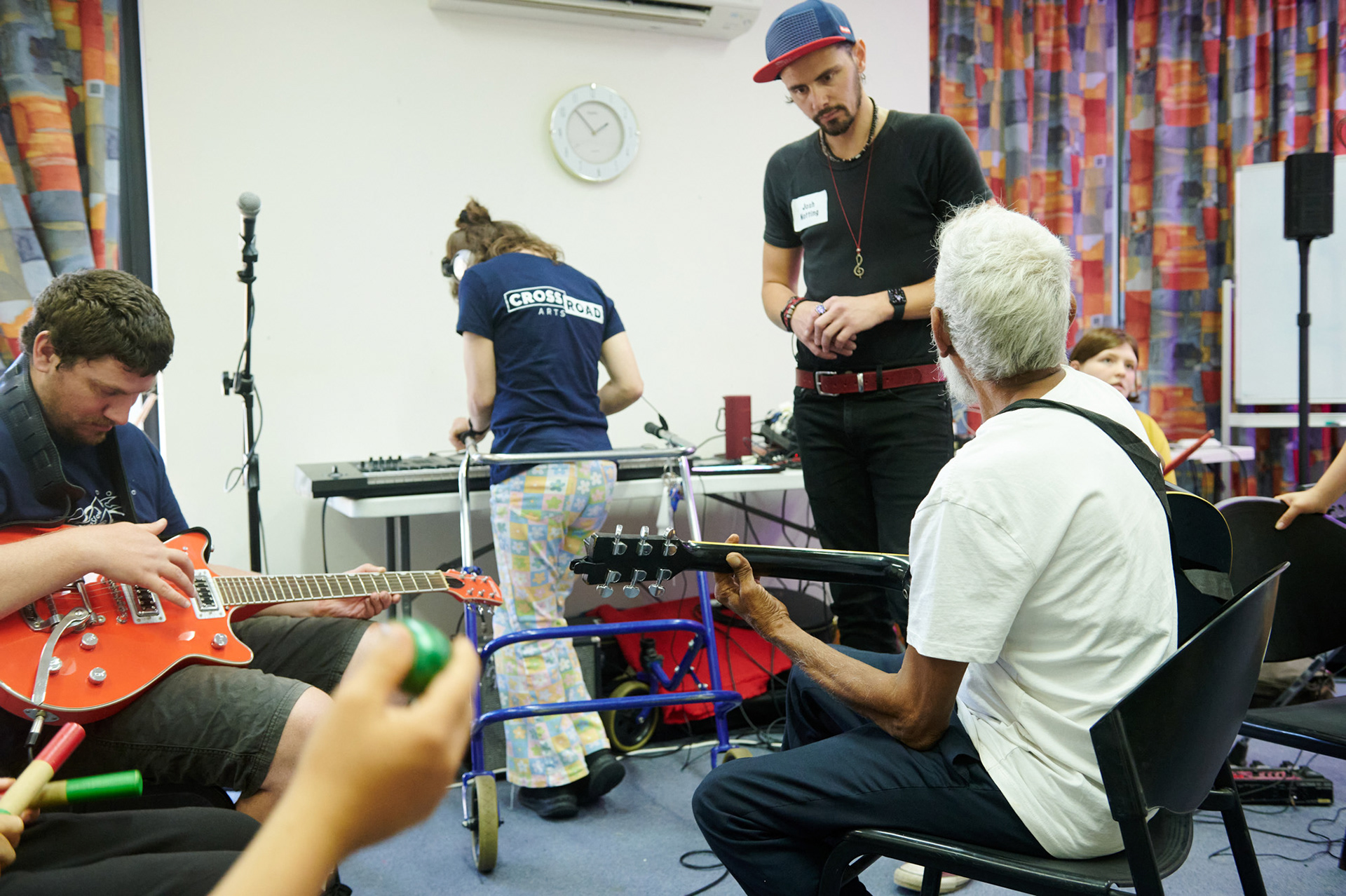 Josh is teaching two men to play the guitar, a lady in the background is playing a keyboard