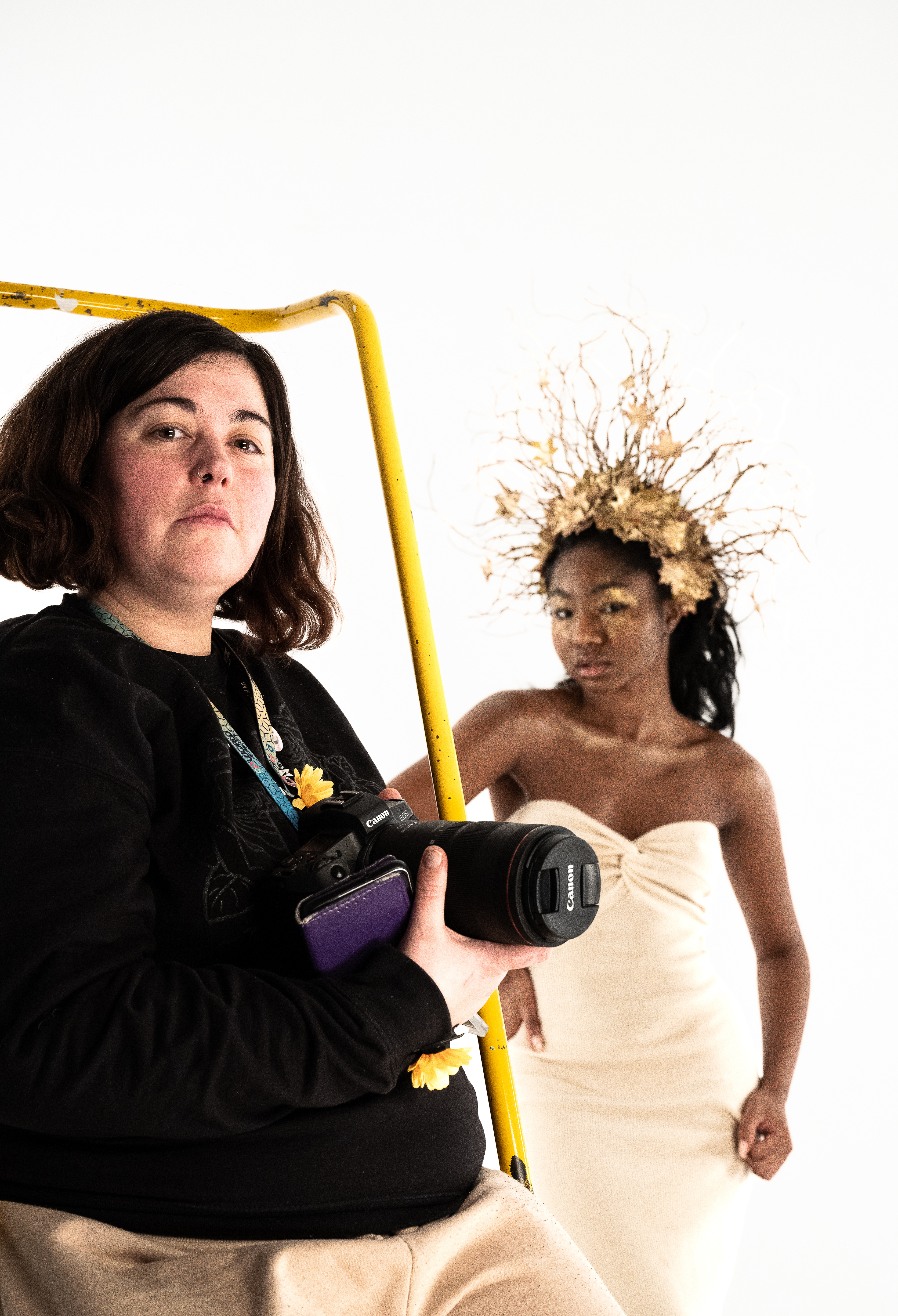 The image is of two women, the first is looking at the photographer, she is dressed in a black top and she has a sunflower pinned to to front. she holds a camera in her hands. the second woman is in the background and is in soft focus , she is wearing a gold head dress made of leaves and sticks .
