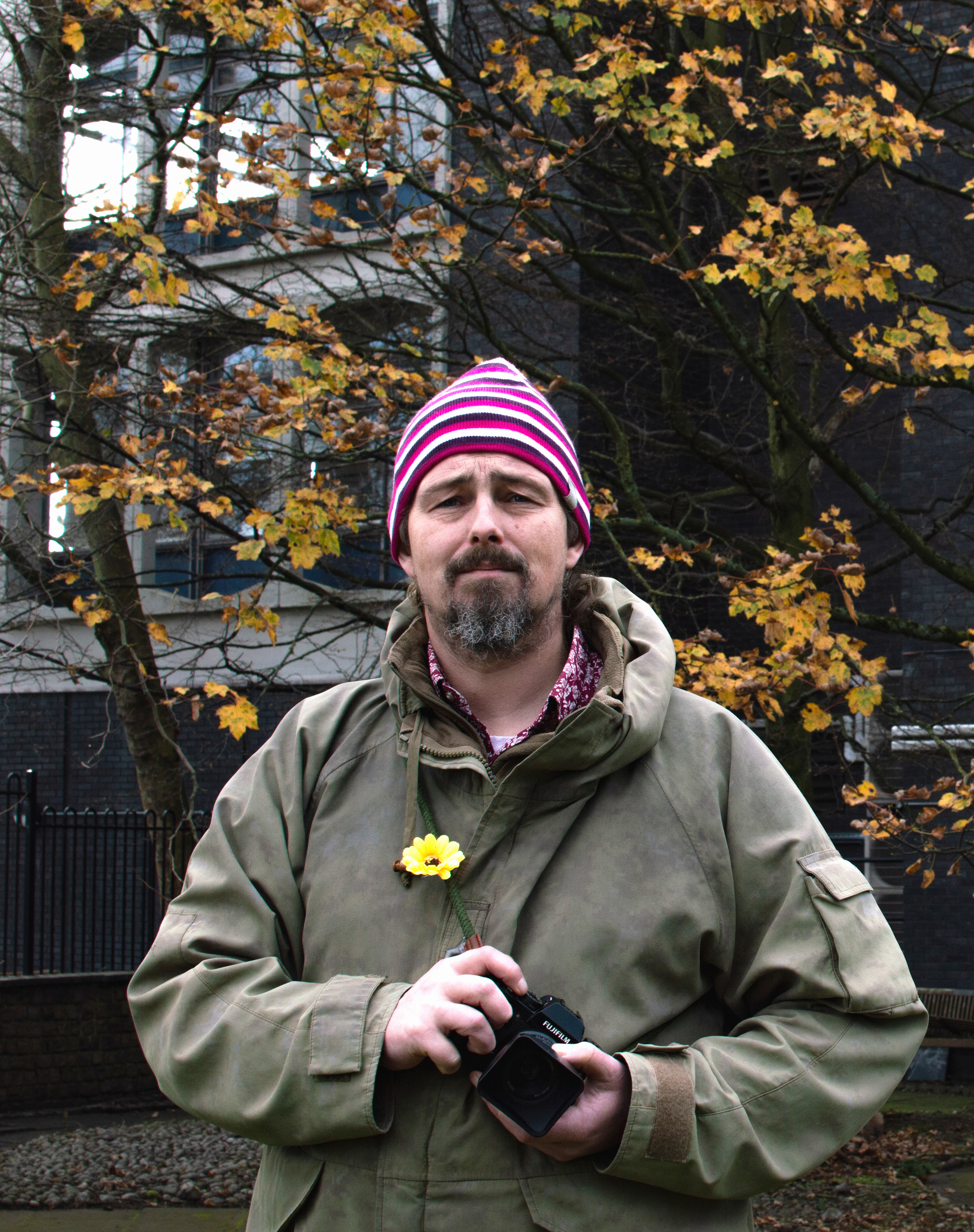 This is a portrait of a man looking towards the photographer, it is shot outside and has a building and trees in the background. The man is holding a camera and has a green jacket on with a sunflower pinned to it. The sunflower represents hidden disabilities